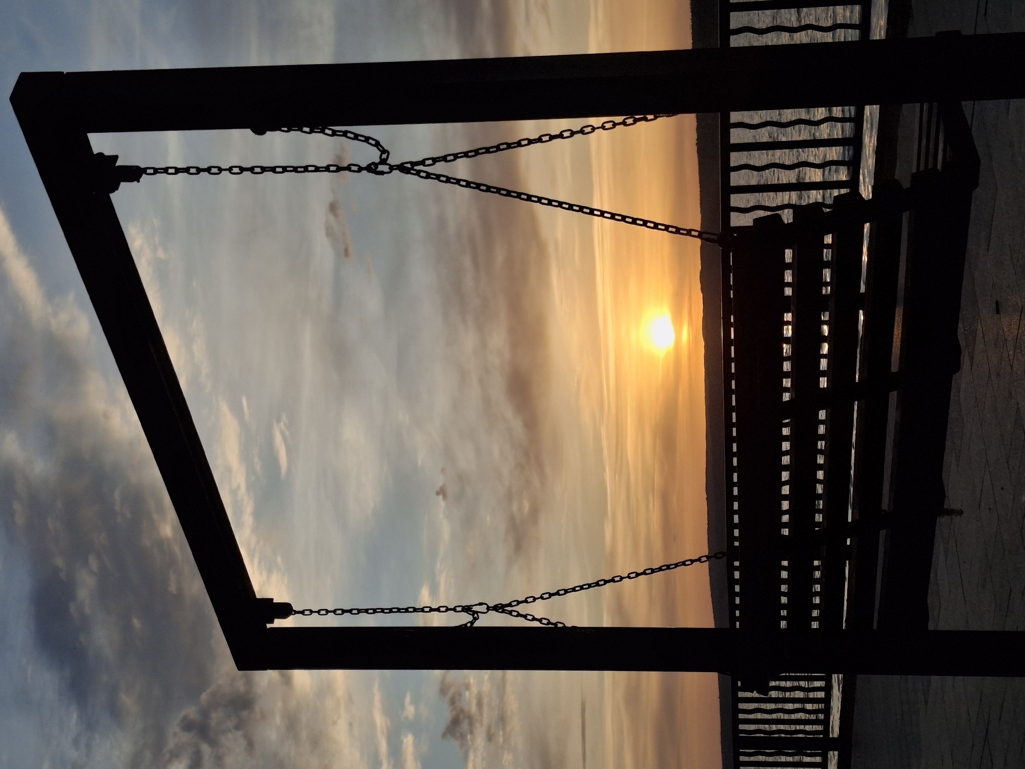 The image depicts a sunset viewed through the frame of a porch swing, likely located on a pier. The swing is silhouetted, suspended by chains. The sun is low on the horizon, obscured slightly by a dark line that could represent a distant shoreline. To the right of the swing is a railing along the pier. The sky is a mix of light blues, oranges, and yellows, indicative of a sunset. Based on the prompt, the location is inferred to be Chaikovsky, Russia.