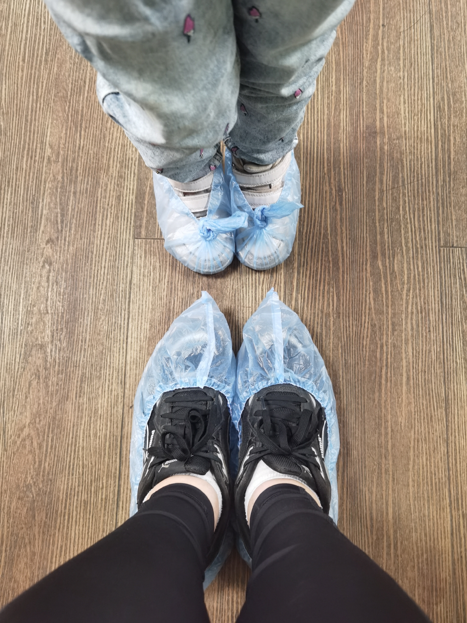 An overhead shot from Izhevsk, Russia, captures the feet of an adult and a child standing on a light brown wooden floor. Both individuals are wearing disposable blue plastic shoe covers over their footwear. In the foreground, the adult's feet are visible, clad in black sneakers (partially revealing the brand "Puma"), white ankle socks, and black leggings. Further back, the child's feet are seen in light-colored sneakers (featuring stripes suggestive of Adidas), light-colored socks, and light blue jeans adorned with small pink ice cream cone patterns. The shoe covers on the child's feet are tied with visible knots over the laces. The wooden floor exhibits a consistent vertical grain pattern. The presence of shoe covers indicates an indoor environment requiring hygiene or protection, such as a medical office, cleanroom, or a private dwelling with strict cleanliness protocols.