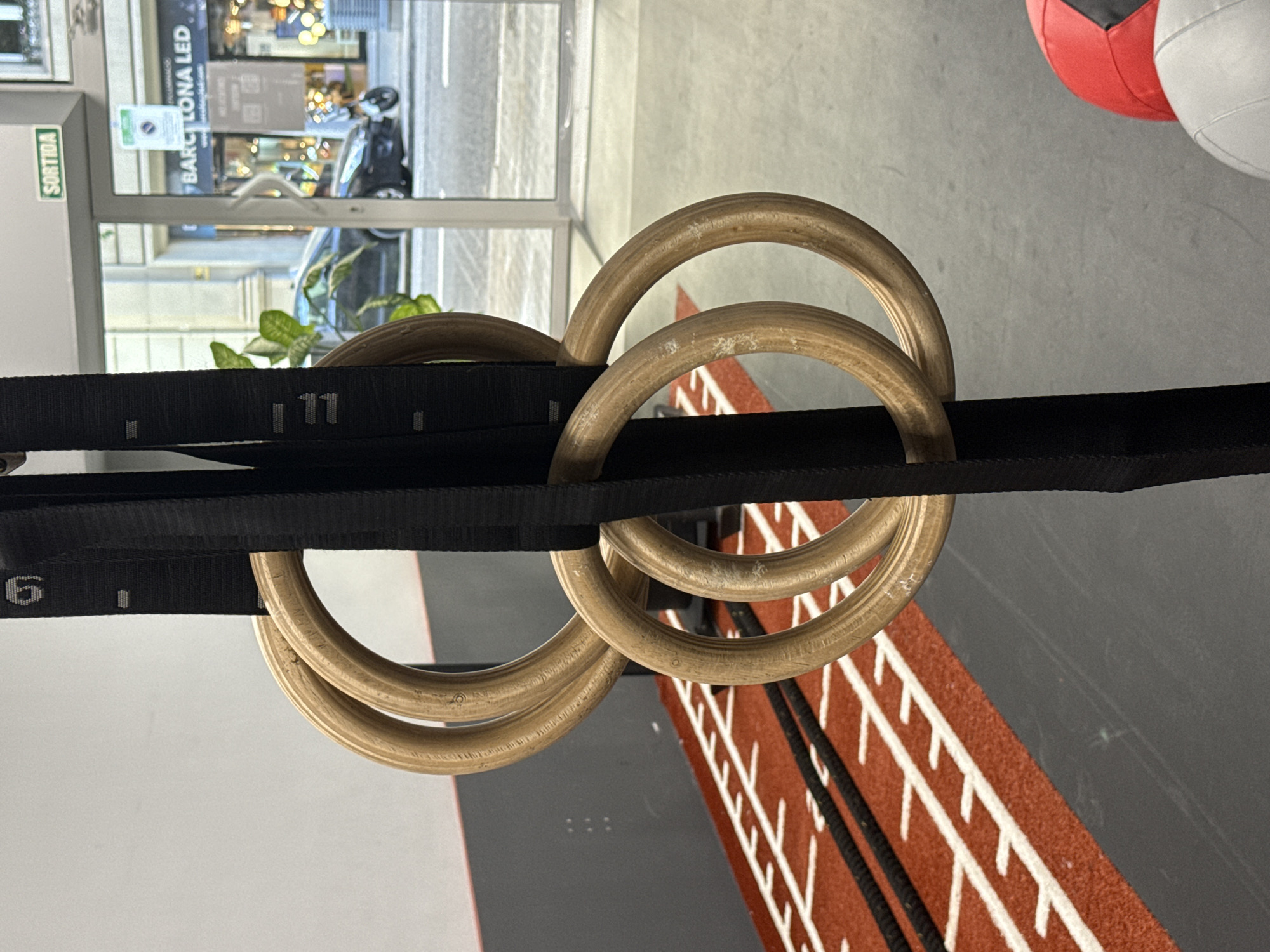 A close-up photograph, originally oriented sideways, captures two sets of wooden gymnastic rings suspended by black straps, indicating a gym or fitness studio environment. The rings exhibit signs of wear, with scuff marks on their light brown wooden surfaces. The black nylon straps are marked with white numbers, including "11" and "6," along with smaller dashes, suggesting adjustable length indicators.
Through a large window or glass door in the background, a bustling urban street scene is visible. Reflections on the glass and blurred architectural details define the street, where a scooter or motorcycle can be vaguely discerned. A significant textual clue, "BARCELONA LED," is visible on a sign across the street, explicitly placing the location in Barcelona, Spain. Near the window, a green sign clearly reads "SORTIDA," which is Catalan for "Exit."
To the right side of the rings, an indoor area is partially seen, featuring the top halves of red and grey medicine balls. Below them, a distinct red floor area with parallel white lines, resembling a running track, transitions into a plain dark grey surface. On the left, a plain white wall and the green leaves of a plant add to the indoor setting. The natural light filtering through the window suggests it is daytime. No people are visible, and no active event is taking place, presenting a static view of fitness equipment within its setting.