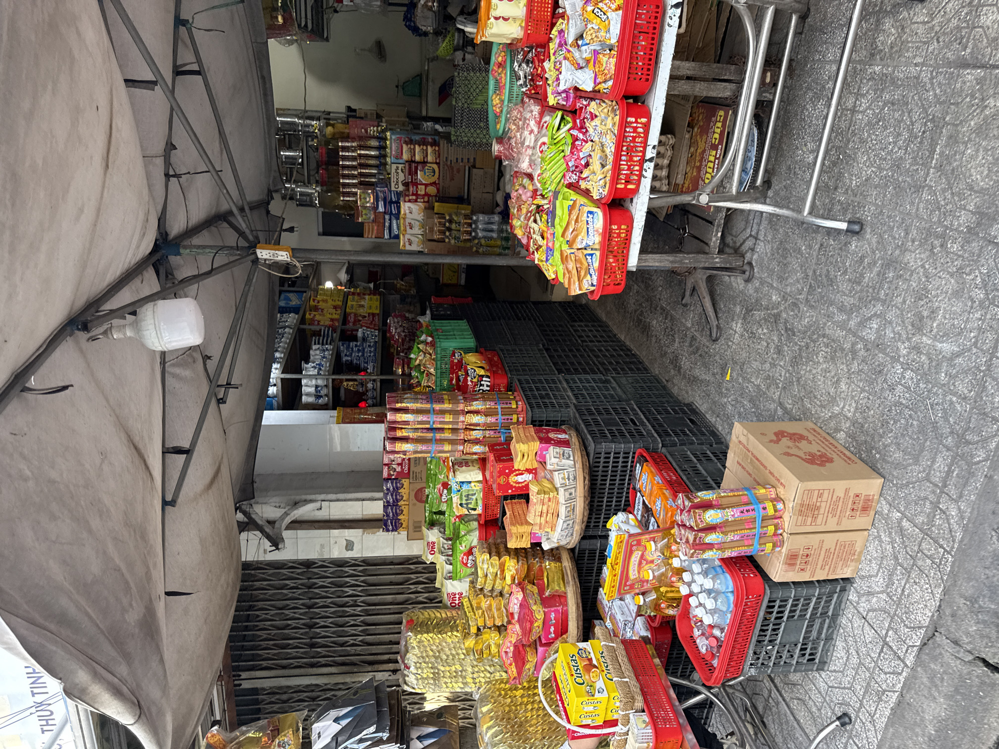 This image depicts a street-level market stall in Nha Trang, Vietnam, likely selling a variety of goods including snacks, incense, and possibly religious offerings. The stall is covered by a tan-colored awning stretched over a metal frame. In the foreground, several red plastic baskets are filled with packaged snacks, such as biscuits and wafers. To the left, yellow boxes labeled "Custas" are visible. Behind these, a collection of gold-colored paper stacks are arranged, possibly for ritual use. A stack of black plastic crates forms a base for displaying some of the merchandise. Further back, more shelves and displays are packed with a diverse array of food items, bottled drinks, and other products. In the background, a metal roll-down gate suggests the entrance to a shop or building. The ground is paved with grey and white square tiles, showing signs of wear and some cracks. The lighting suggests it is daytime, with natural light filtering through the awning. No people are clearly visible in the frame. The overall impression is that of a bustling, densely stocked market environment.