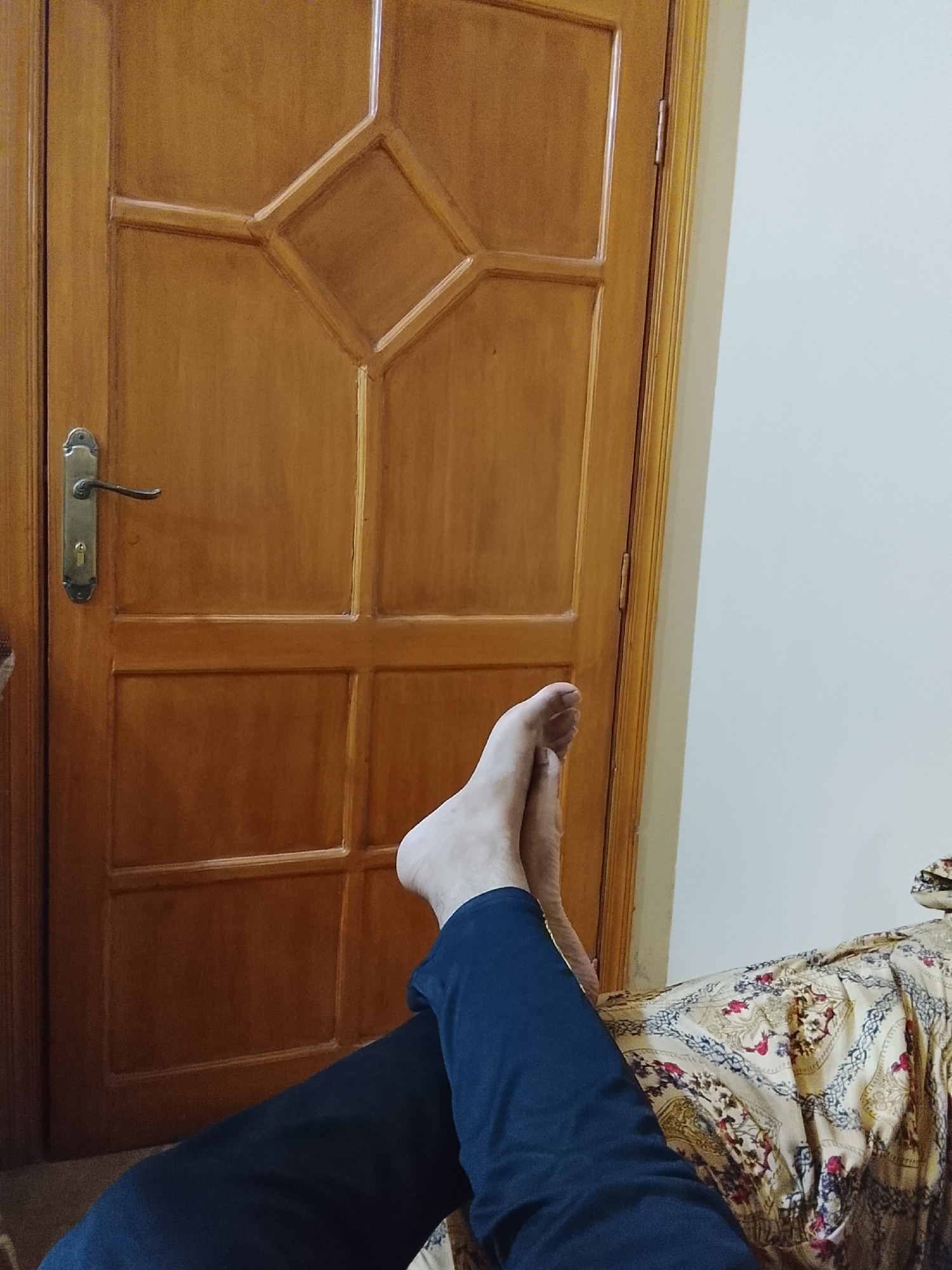 The image shows a partial view of a person reclining. Two bare feet are visible, with the lower legs covered in blue pants extending toward the bottom of the frame. The person is lying on a patterned bedspread. Behind the person, there is a wooden door with a diamond-shaped design and a silver handle, and a plain white wall. The location is presumed to be a private indoor space. The city is Multan, Pakistan.