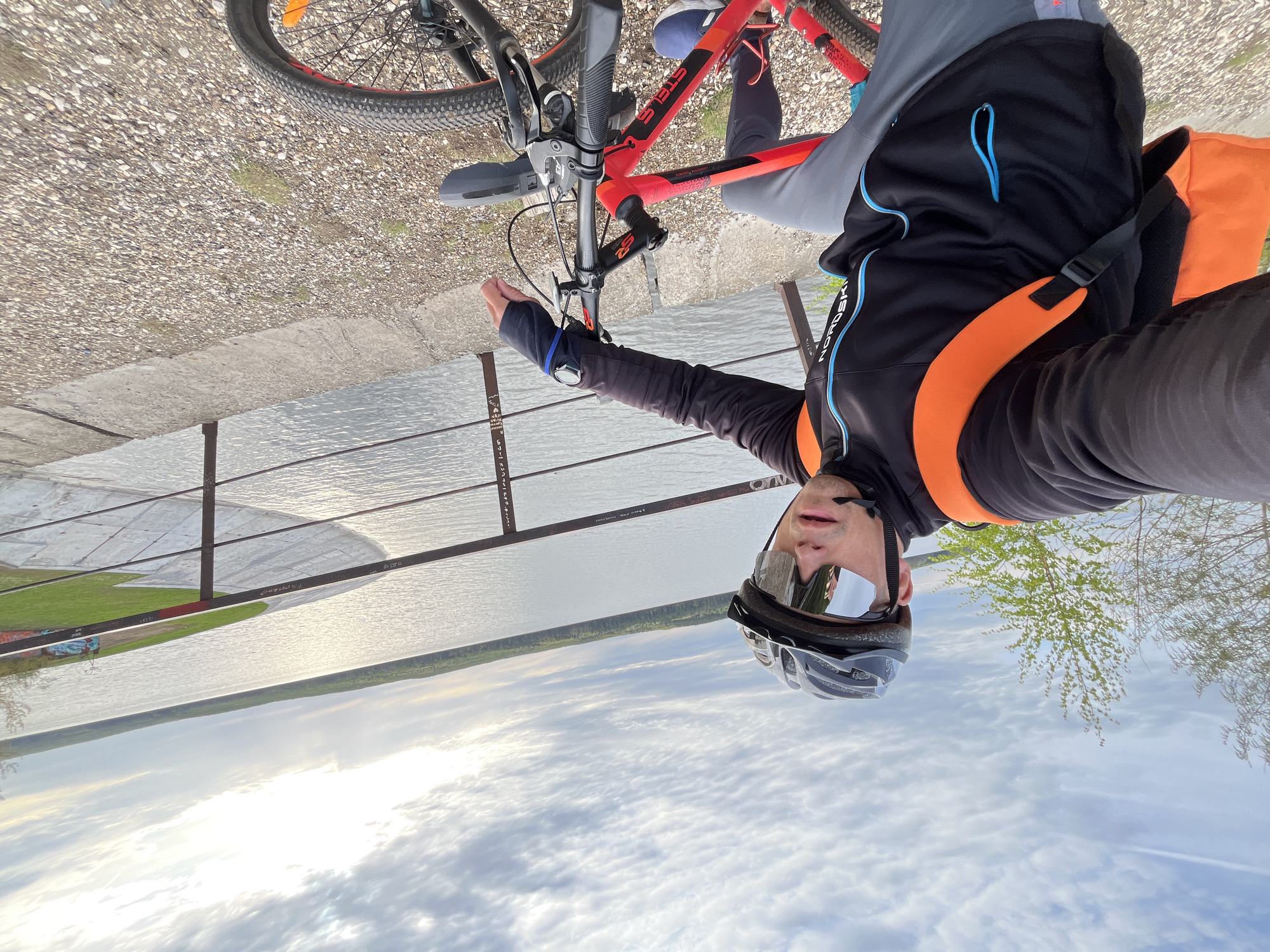 The image shows a selfie of a person wearing a black helmet with sunglasses and a black and orange jacket, posing with a red bicycle. The person is standing on a walkway with a metal railing overlooking a body of water, likely a river or lake. Across the water is a treeline. The sky is visible with clouds. The location is identifiable as Chaikovsky, Russia based on information on the barrier in the background.