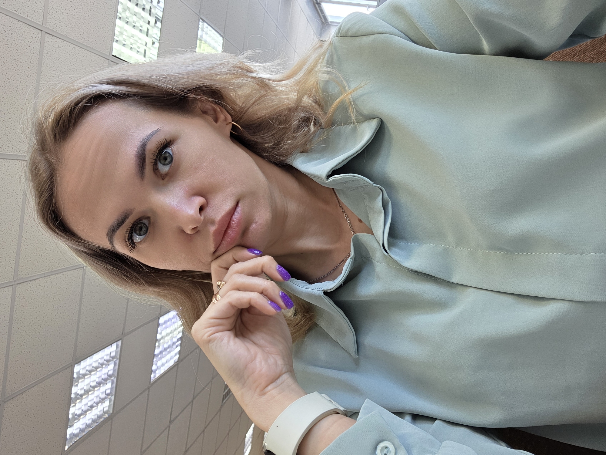 The image depicts a female subject indoors, located in the city of Chaikovsky, Russia. The Caucasian female, with blonde hair and light eyes, is looking directly forward with a neutral expression. She is wearing a light mint-green collared shirt, a thin gold chain necklace, and small gold hoop earrings. Her right hand, featuring purple glitter nail polish, two gold rings, and a white wristband, rests with fingers against her chin and jawline. The background comprises a white suspended ceiling with a grid pattern and multiple recessed rectangular fluorescent light fixtures. A bright window or external light source is partially visible on the right side.