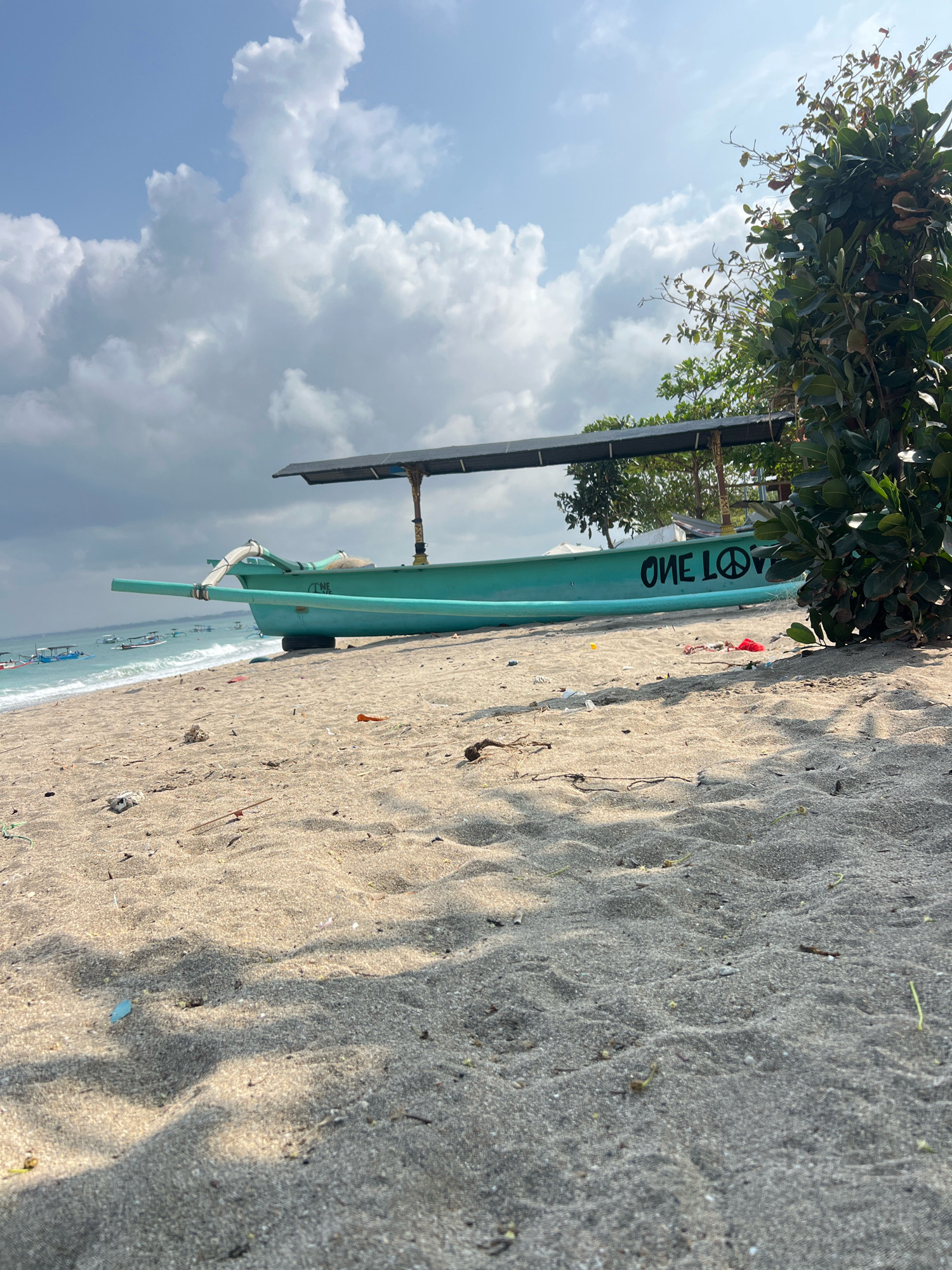 A turquoise outrigger boat rests on the sandy shore of Kuta Beach, Indonesia. The boat, with a covered seating area, displays the black lettering "ONE LOVE" and a peace symbol on its side. Scattered debris, including plastic wrappers and a red piece of fabric, litters the sand in front of the boat. In the distance, the turquoise ocean meets a hazy sky filled with dramatic, white clouds. The foreground is dominated by the textured sand, with shadows cast by unseen foliage to the right. Several other similar boats are visible further down the beach and on the water.