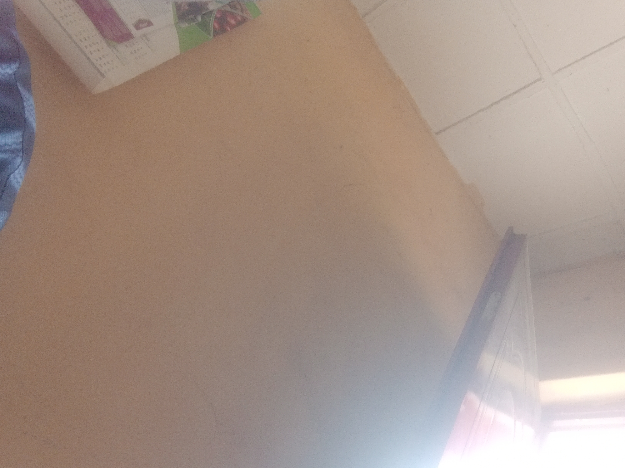 The image shows a close-up, angled view of a wall and ceiling. The wall is painted a light, peachy-beige color. The ceiling appears to be made of white tiles with visible seams. In the upper left corner of the image, a portion of what looks like a calendar or poster is visible, with text and some colored images. A blue fabric item is also partially visible on the left edge. On the right side of the image, a white door frame with some decorative molding is angled towards the viewer, with a bright light source emanating from the right, suggesting a window or open doorway. The overall impression is of an indoor domestic or office space. This scene is not specific to Jalingo, Nigeria.