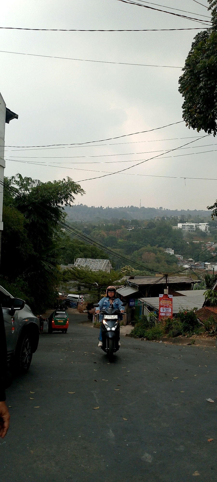 The image shows a person riding a scooter down a paved, uphill street in what appears to be a residential area of Bandung, Indonesia. The rider, wearing a denim jacket and a helmet, is facing forward. On the left side of the street, a silver car is partially visible. In the background, there are houses and buildings nestled among lush green trees and rolling hills, characteristic of Bandung's hilly terrain. Overhead, power lines crisscross the sky, and the sky is overcast. A green and orange vehicle is parked further up the street, and a red and white banner is displayed on a pole on the right side of the road. The scene suggests a typical day in a densely populated but verdant city.
