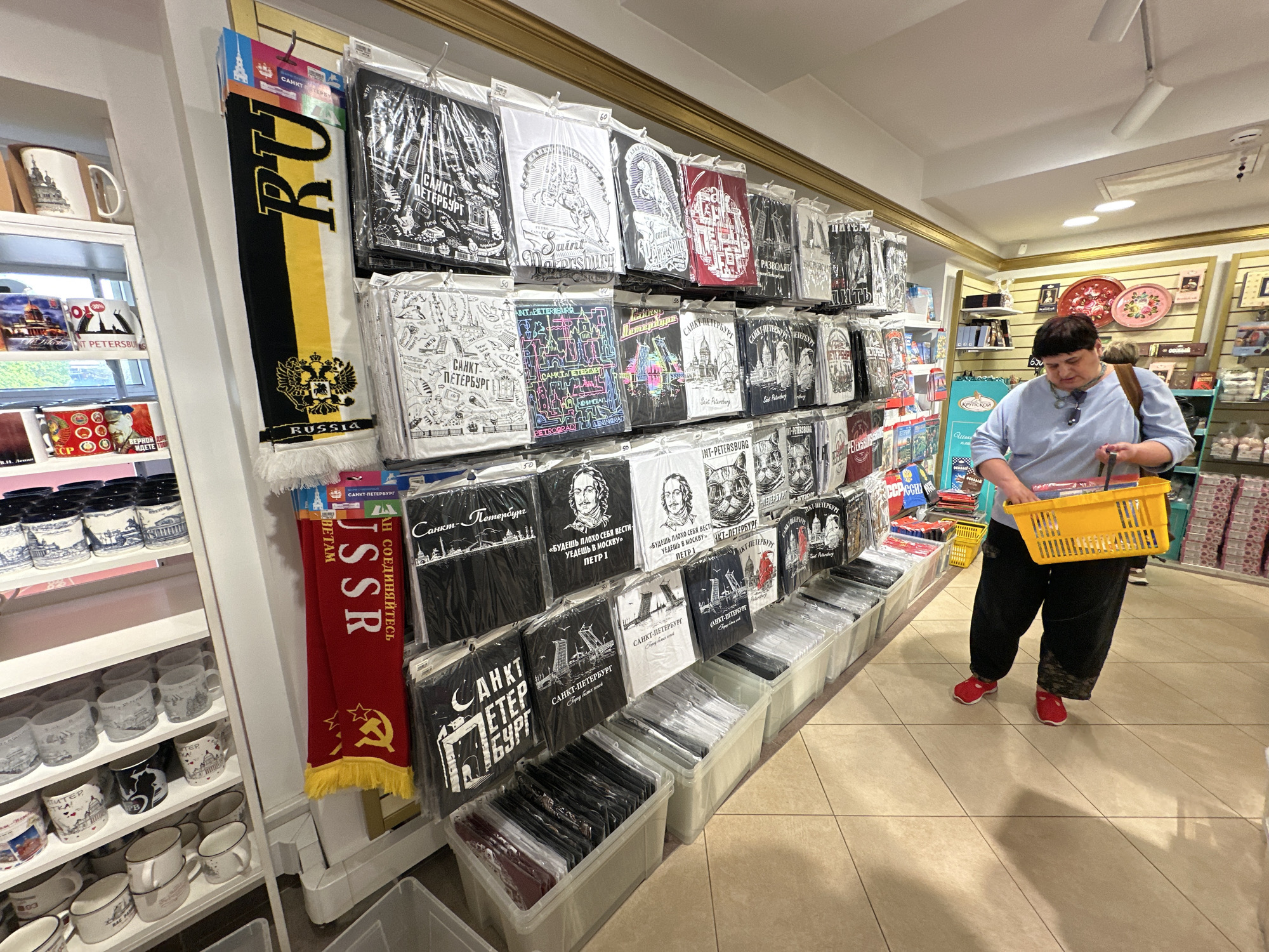 A well-lit retail space displays a variety of souvenirs, particularly clothing items and mugs, likely in Saint Petersburg, Russia, given the Cyrillic inscriptions on the merchandise. A woman with short, dark hair wearing a light blue top, dark pants, and red sneakers is holding a yellow shopping basket and appears to be shopping. Souvenirs such as t-shirts, mugs, and scarves line the shelves and walls, bearing images and text related to Saint Petersburg. The store seems to cater to tourists, offering items with Russian iconography and text, along with images of historical figures like Peter the Great. The environment is a typical souvenir shop setting.