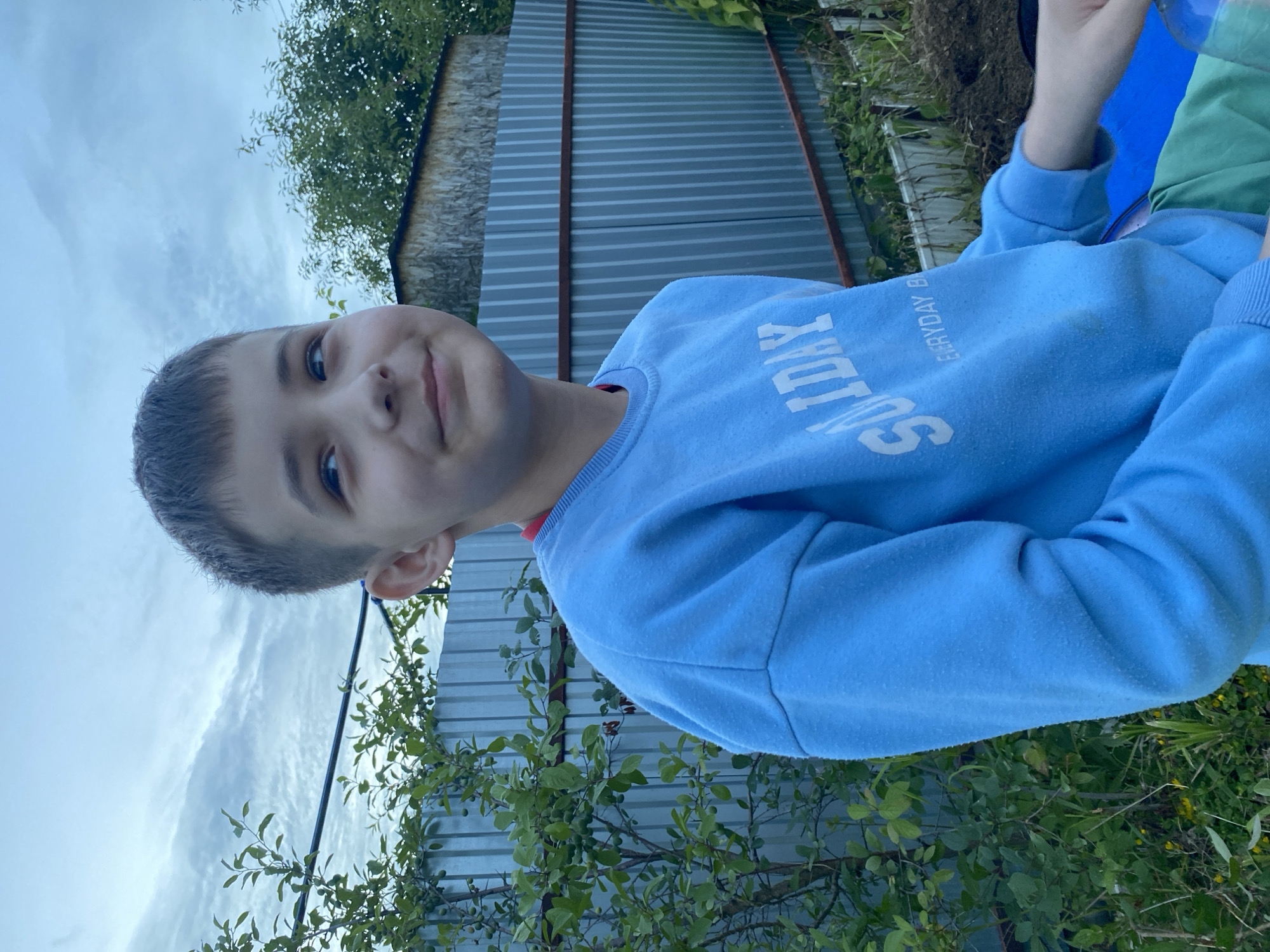 The image shows a close-up of a young boy wearing a light blue sweatshirt with the text "SUNDAY" printed on it in white lettering. He is looking at the camera with a slight smile. The background is outdoors, with a corrugated metal fence and greenery visible. The sky is overcast, suggesting it is daytime. The boy's hair is short and dark. The sweatshirt appears slightly pilled. The overall impression is casual and candid. The image appears to be taken in Chaikovsky, Russia.