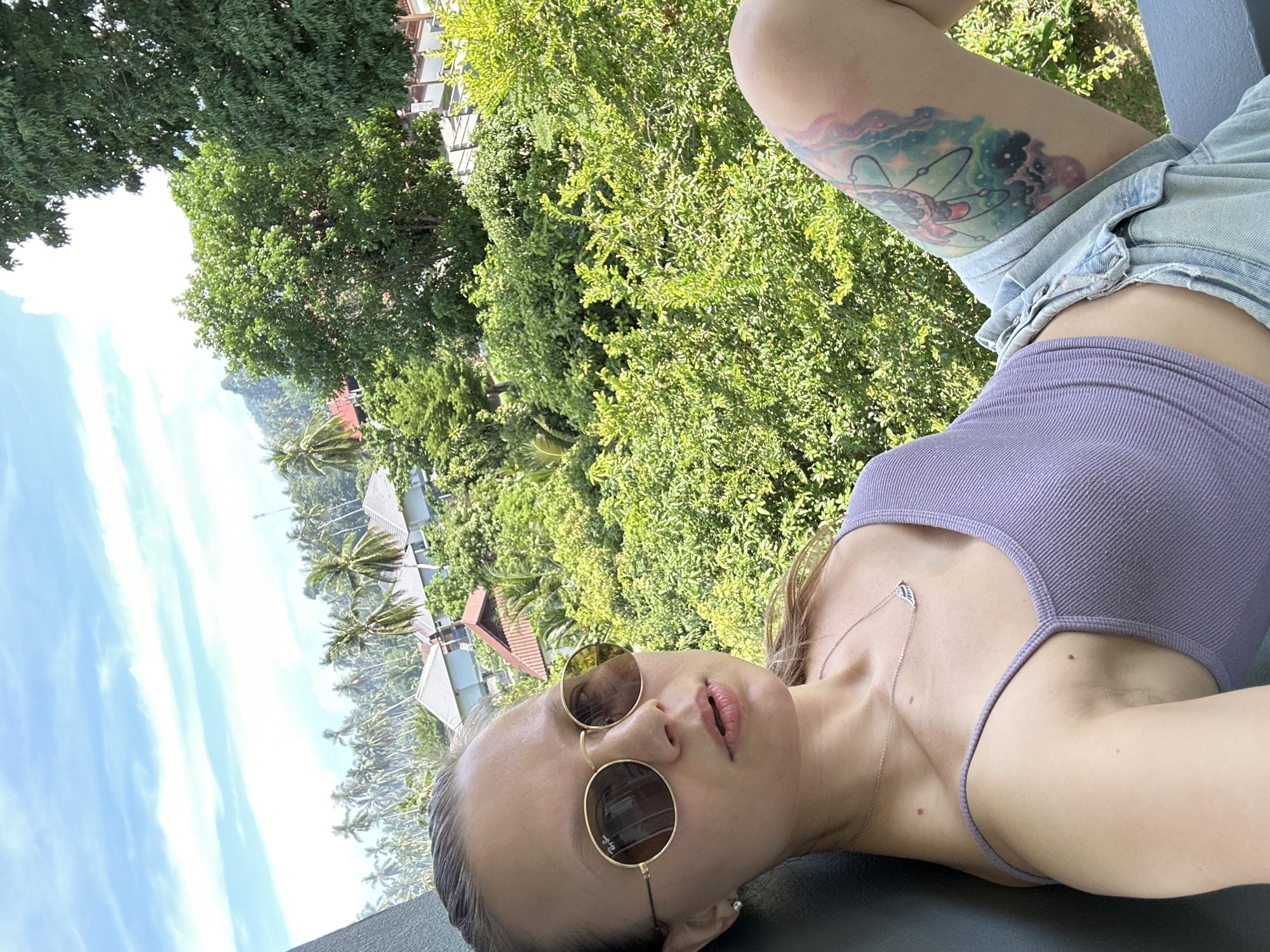 A woman is taking a selfie outdoors with lush green foliage and palm trees in the background. She is wearing round, gold-rimmed sunglasses, a light purple tank top, and denim shorts. A tattoo is visible on her left thigh. In the distance, a cluster of buildings with terracotta roofs can be seen amidst the trees. The sky is blue with scattered white clouds. The scene appears to be a resort or residential area in a tropical location, likely Baan Hua Thanon, Thailand.
