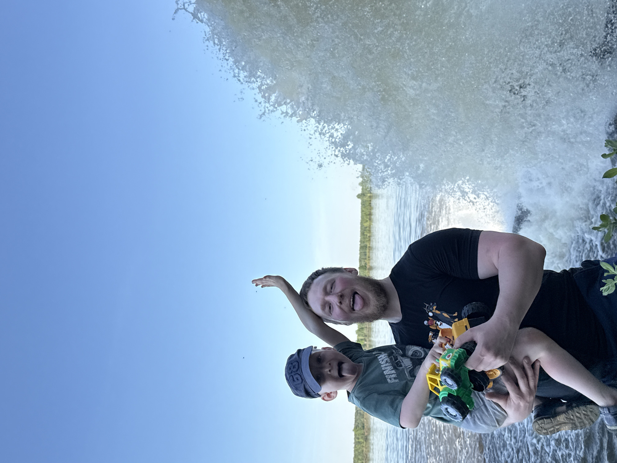 A man and a child stand near a waterfall. The man, with a beard and black t-shirt, holds a toy tractor. The child, wearing a cap, is held in the man's arms. They are both sticking their tongues out. The background includes a body of water and vegetation. The location may be in Novyi, Russia, as specified.