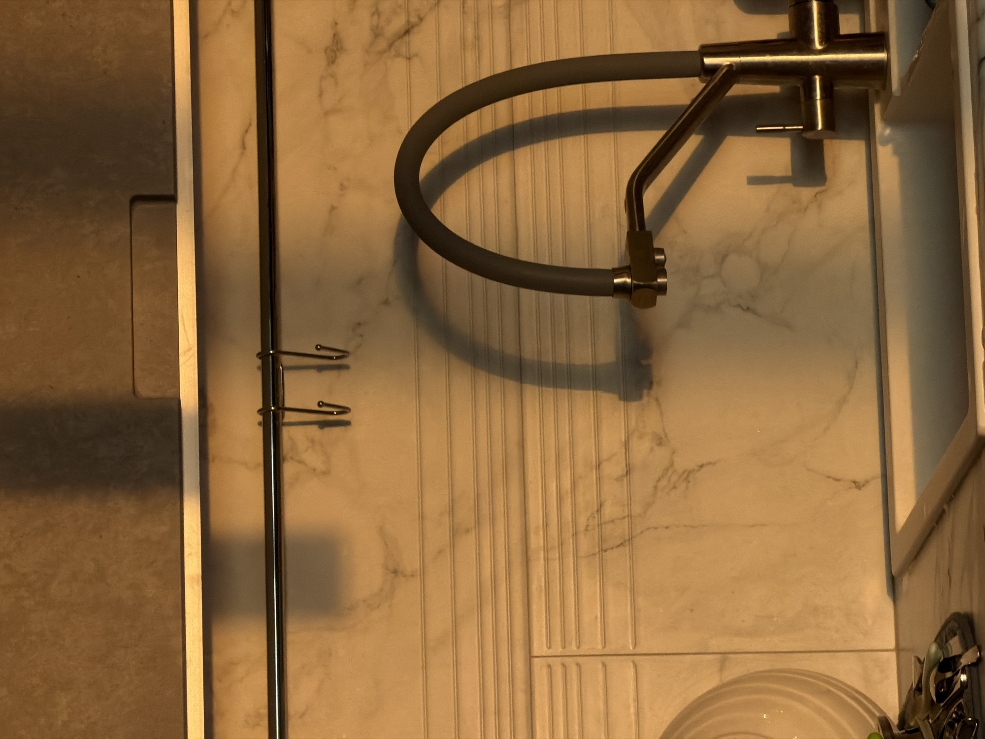 The image shows an elevated view of a bathroom sink and faucet. The sink has a white marble-like surface with vertical grooved patterns. A gray flexible hose curves over the sink surface, connected to a chrome faucet attached to the wall. A chrome towel rack with two hooks is mounted vertically on the left. The overall lighting appears warm, possibly from artificial sources. A portion of the countertop and sink basin is visible in the bottom right corner. The environment is clean and modern.