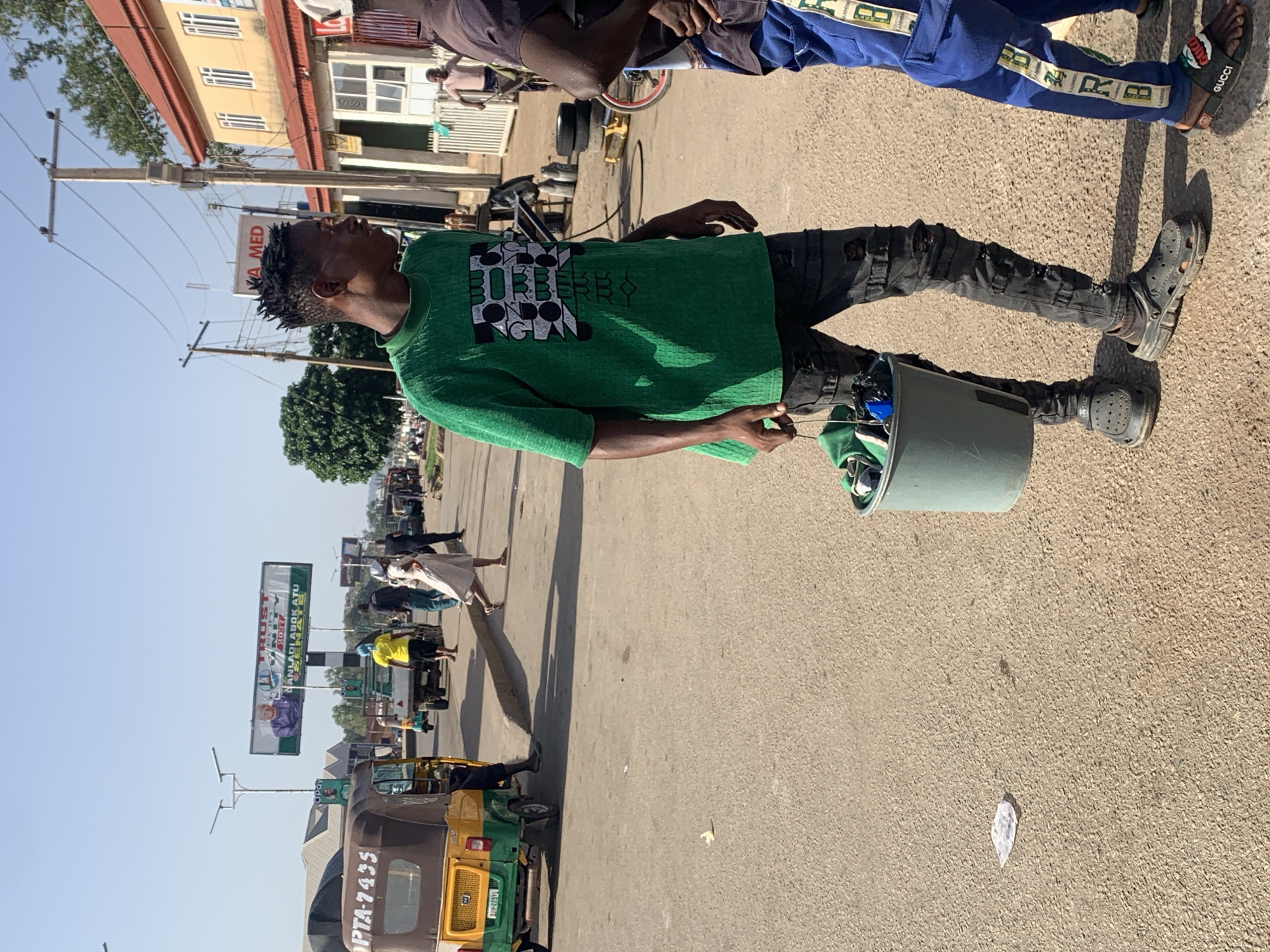 A daytime outdoor shot depicts a street scene in Gura Top, Nigeria. A young man wearing a green Burberry-logo T-shirt, ripped black jeans, and black Crocs is standing on the pavement, holding a grey bucket containing various items. Another person, partly visible, wears blue pants with white stripes and Gucci sandals. In the background, a road features auto rickshaws, pedestrians, and a billboard advertising "Trust...2023." Buildings, power lines, and trees are also visible. The weather appears sunny with a clear sky.