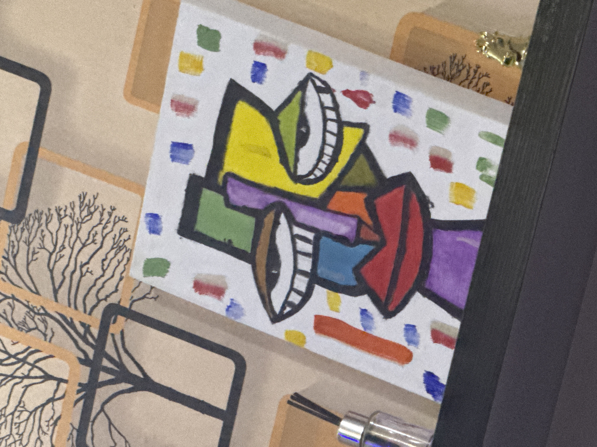 The image shows a close-up of a colorful, abstract painting on a white canvas. The painting appears to depict two faces with cartoonish, exaggerated features, including large eyes with thick lashes. The faces are composed of various geometric shapes and colors such as yellow, green, purple, red, and blue, outlined in black. The background of the canvas is dotted with small squares of color. The painting is set against a beige surface decorated with stylized images of trees or branches with a black outline. In the upper-right corner, there is a small section of decorative gold foliage. In the lower portion, a part of a silver-colored cylindrical object, potentially a pen or cleaning tool, is visible. A dark, solid-colored object is partially obscuring the right side of the canvas. There is no indication in the image to suggest that it is located in Jos, Nigeria.