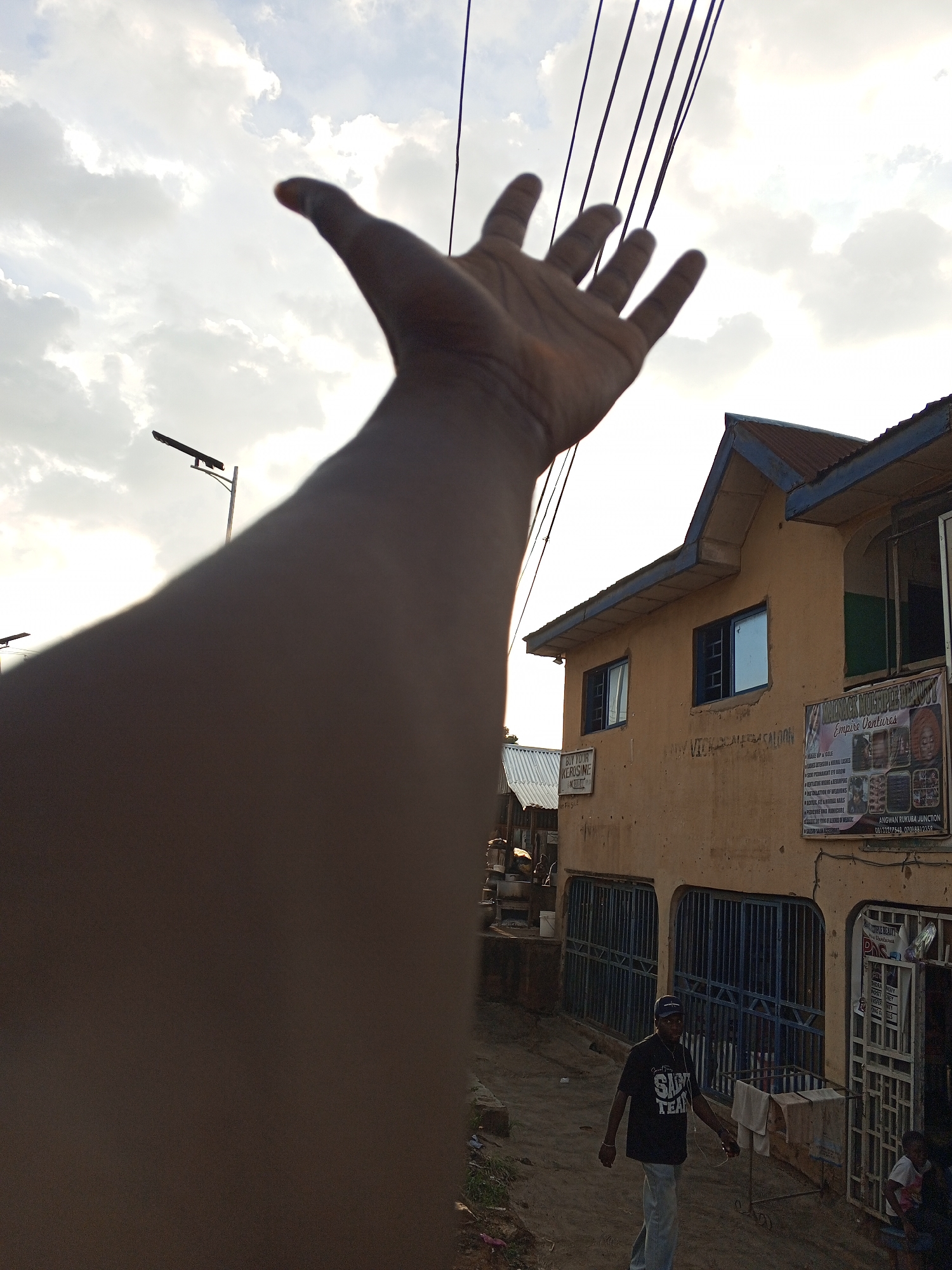 A man's arm is extended towards the sky with an open hand. Overhead electrical wires are visible. In the background, there is a two-story, light-brown building with blue-trimmed windows and a rust-colored roof. Signs on the building's wall advertise "KEROSINE" and "MINACK MULTIPLE BEAUTY Empire Ventures" with contact information.
A man wearing a black "SAINT TEAM" t-shirt and light-colored pants walks on a dirt road. Nearby there is a metal gate, what looks like a stall, and a child standing in a doorway. The location appears to be a street in Gwong, Nigeria. The weather is slightly overcast with bright sky.