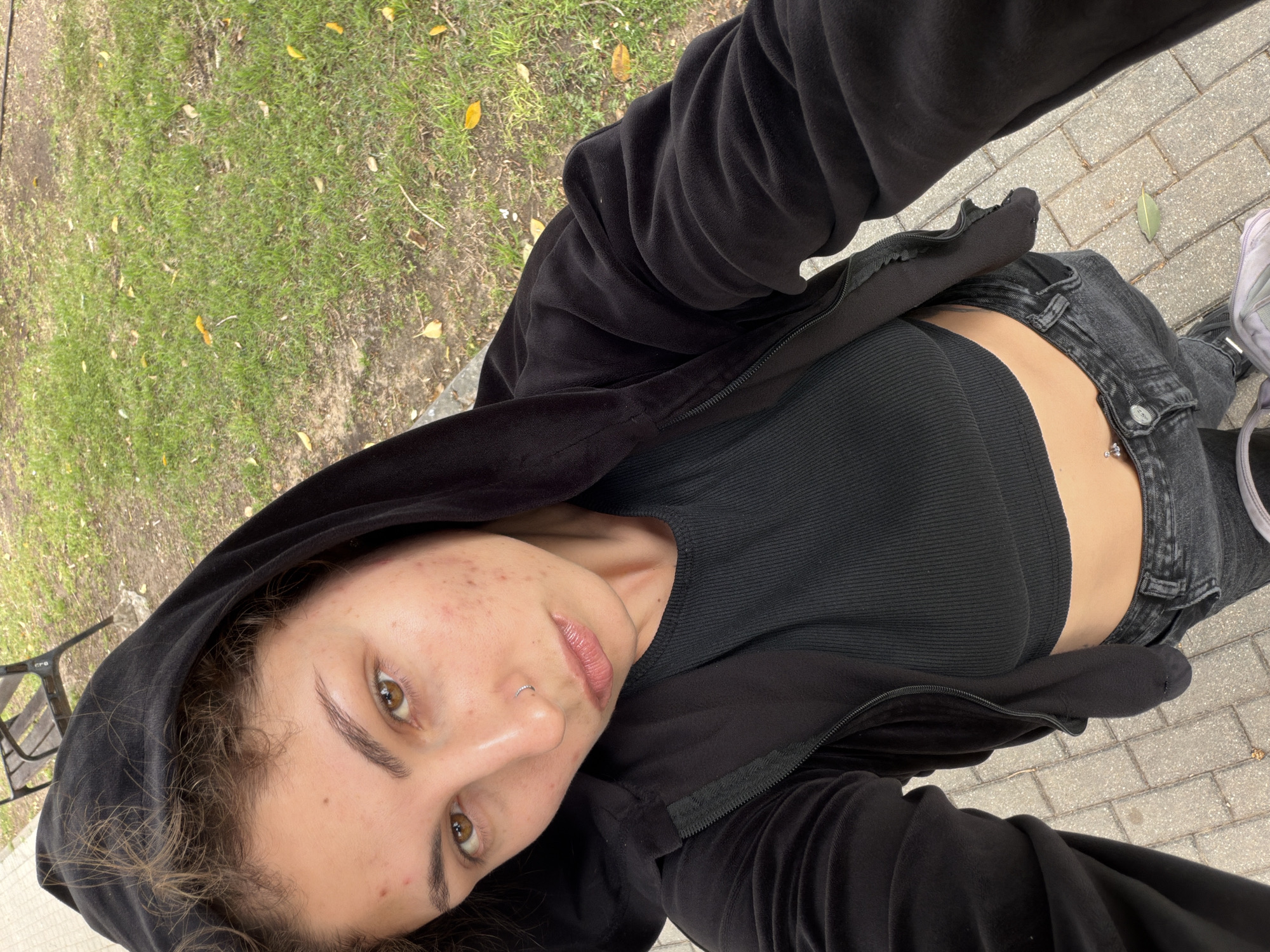 A young woman is taking a selfie outdoors. She is wearing a black hooded jacket, a black crop top, and grey jeans. Her skin tone is fair with freckles. The background consists of green grass and brick pavement. The location is inferred to be Kiryat Bialik, Israel.