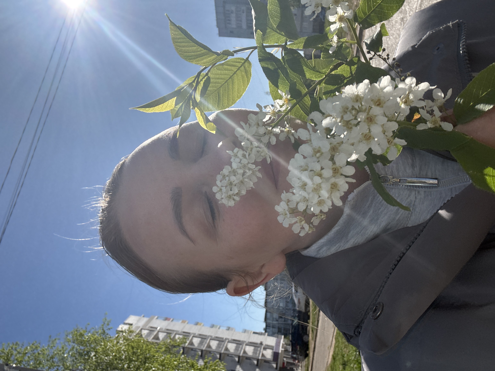 A person is holding a branch of white flowers near their face. The person has fair skin, closed eyes, and light brown hair. They are wearing a gray jacket over a lighter gray top. The background shows a blue sky with the sun shining brightly, partially obscured by the flowers. Buildings and trees are visible in the lower part of the frame, suggesting an urban environment. The location is the city of Chaikovsky, Russia.