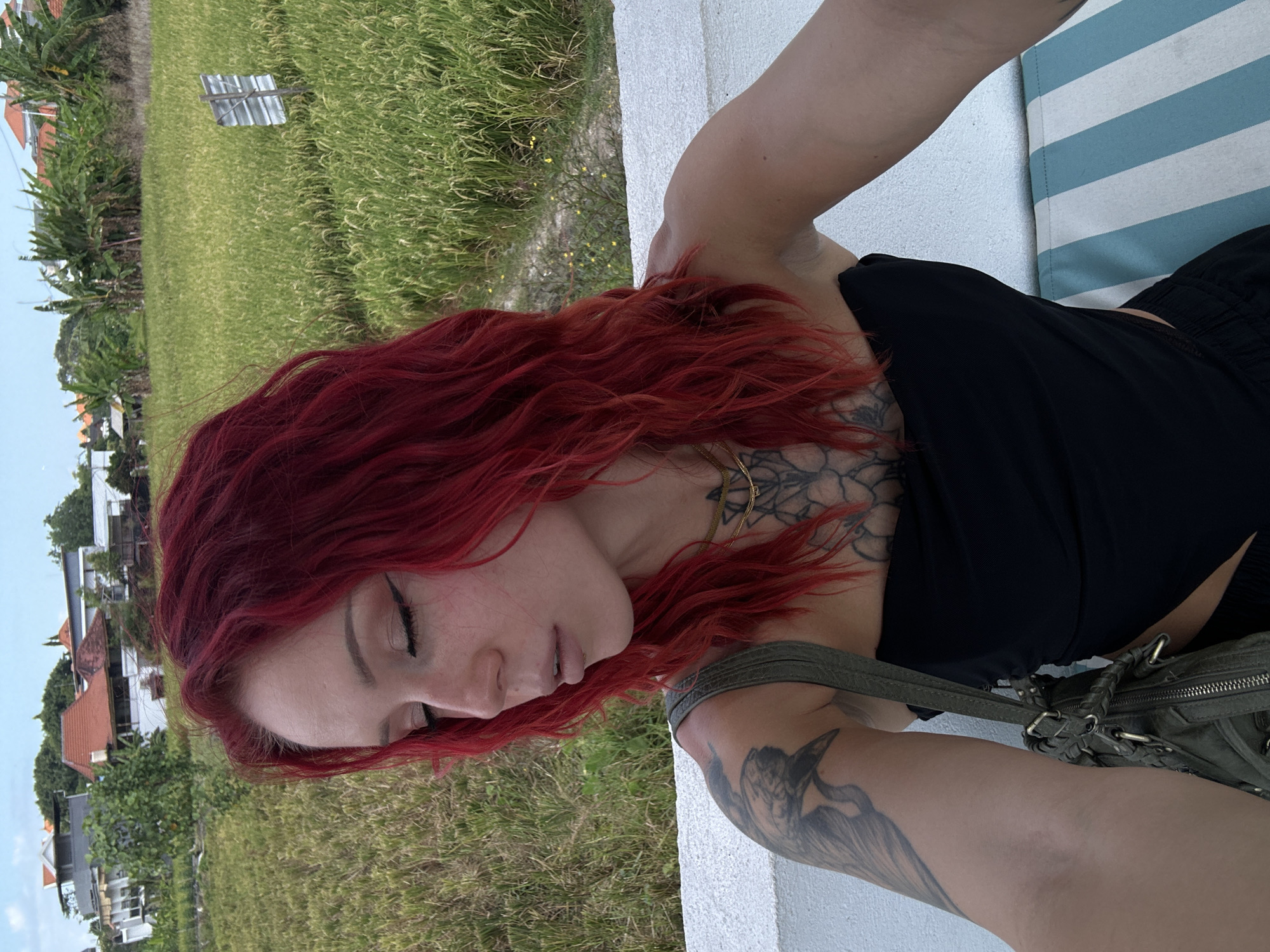 A woman with vibrant red hair is pictured in a close-up selfie. She is wearing a black strapless top and a gold necklace. Tattoos are visible on her left arm and chest. She is holding a green, woven handbag on her left shoulder. In the background, there is a lush green rice paddy field, some buildings with red roofs, and palm trees under a partly cloudy sky. The scene appears to be in North Kuta, Indonesia. The woman is looking down with her eyes closed, and her makeup includes winged eyeliner and neutral lipstick. She is sitting on a white ledge, with a striped cushion visible in the lower right corner.