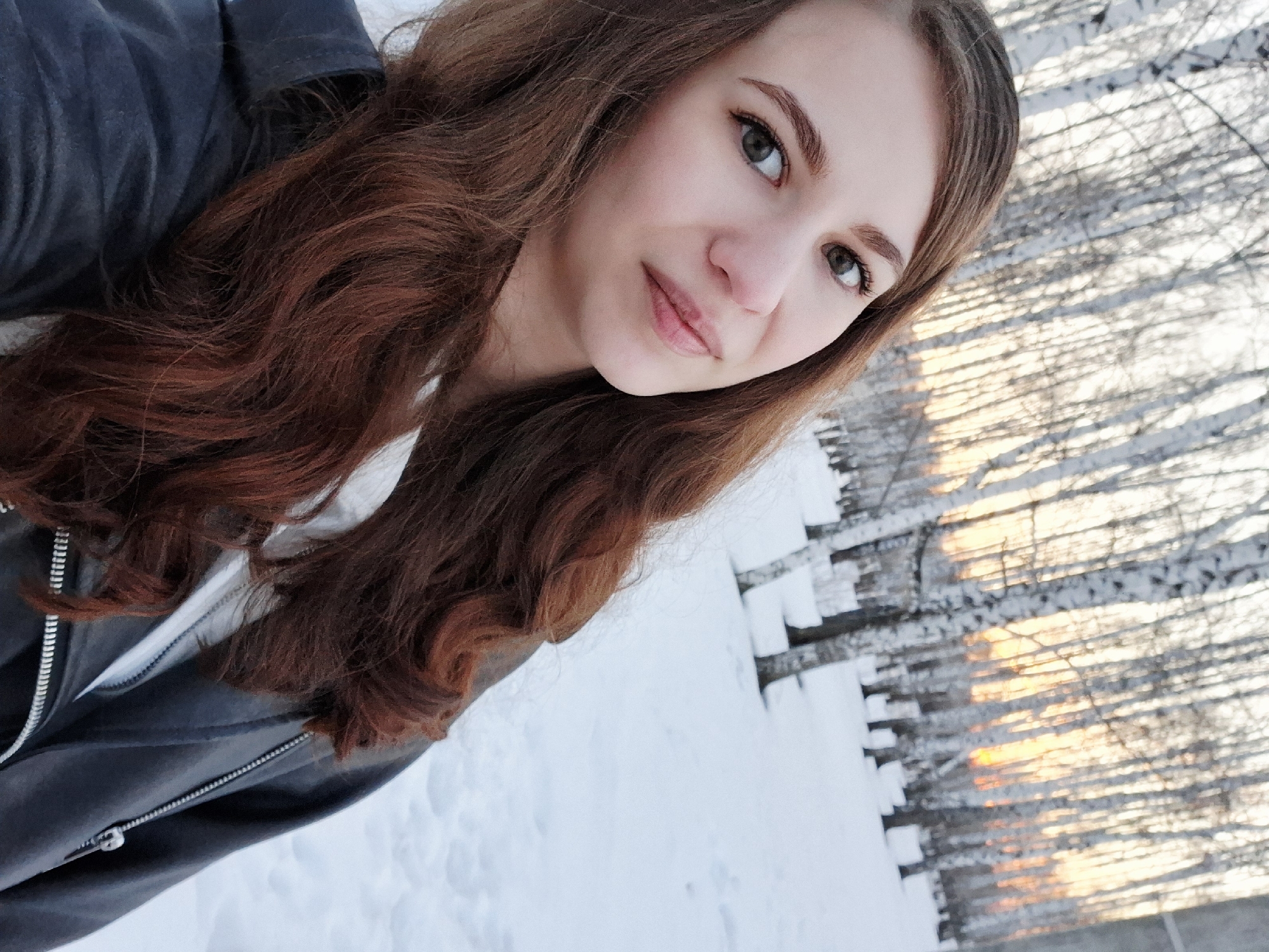 A young woman poses for a selfie in a snowy landscape. She has long, wavy auburn hair and is wearing a dark grey jacket with a white hooded sweatshirt underneath. The background reveals a winter scene with snow-covered ground and a dense thicket of bare trees, likely birch trees based on the white bark and dark markings. The setting sun is visible through the trees. The location is likely in or around the city of Chaikovsky, Russia.