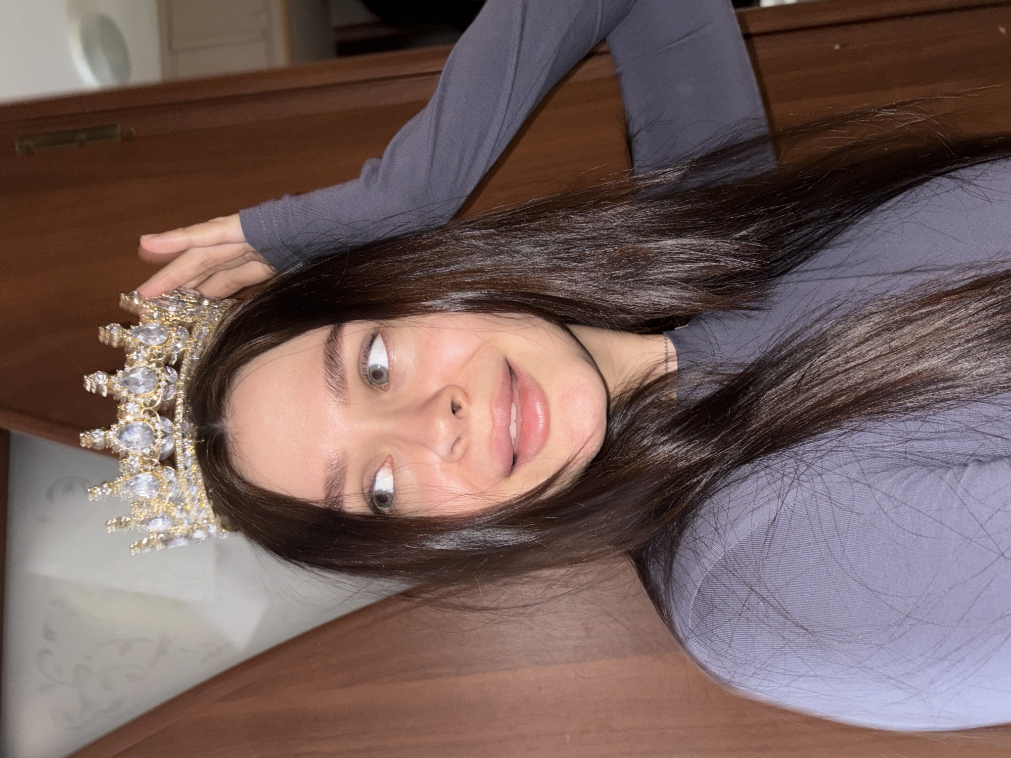A woman is lying on a brown surface, wearing a long-sleeved grey shirt and a jeweled crown. Her dark hair is spread around her. Her eyes are light-colored. Her mouth is slightly open. The photo appears to be taken indoors. The city of Chaikovsky, Russia is not detectable in the image.