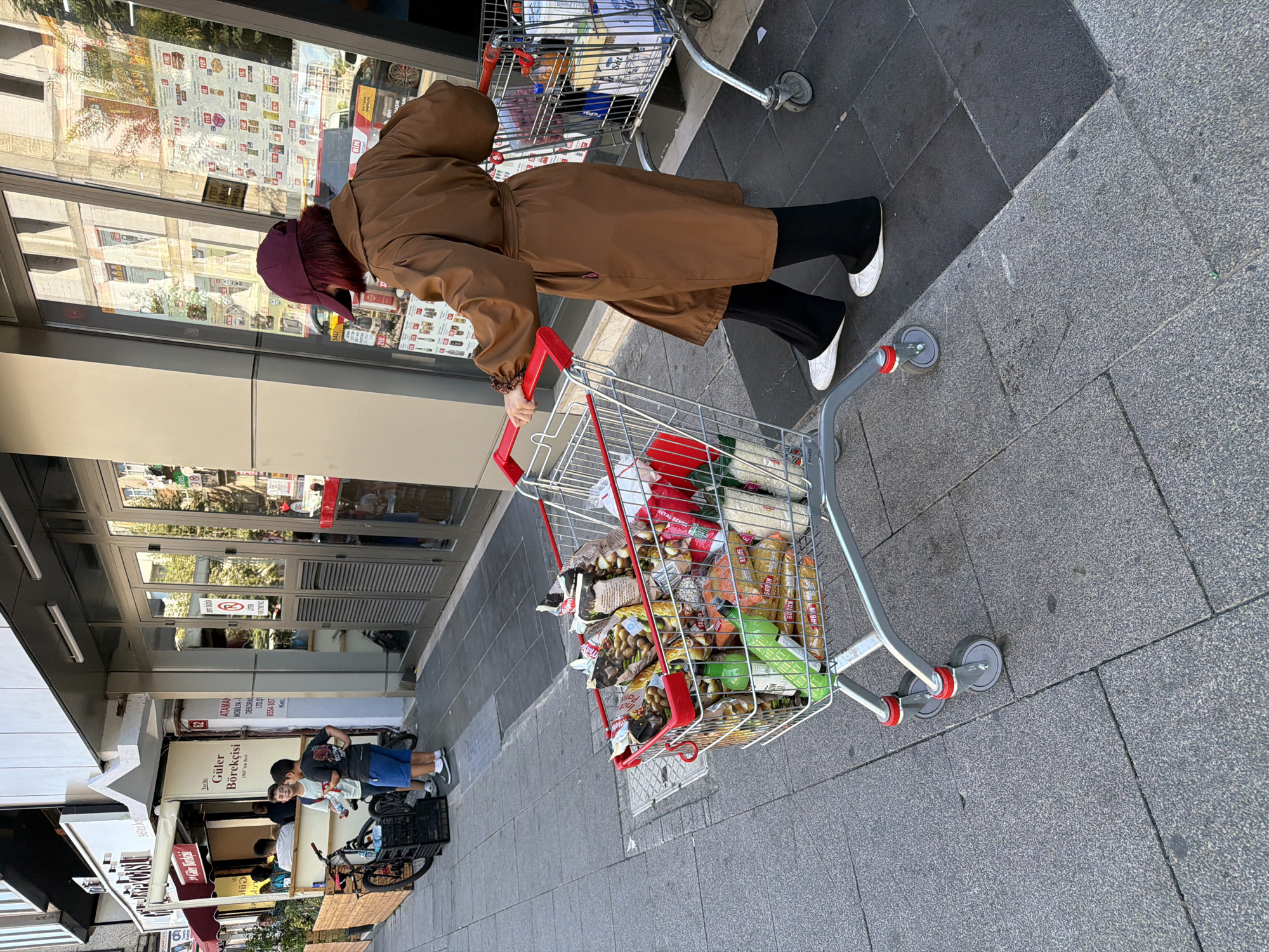 A woman wearing a brown trench coat, black pants, white slip-on shoes, and a maroon baseball cap is pushing a full shopping cart on a sidewalk in Kadıköy, Turkey. The shopping cart is filled with various grocery items, including bags of snacks, bottles, and boxes. Behind her, two young boys stand near a bicycle with a black basket. They are wearing casual clothing. To the left of the scene, there is a storefront with outdoor seating, and to the right, a large glass-fronted building with signs and advertisements. The sidewalk is paved with grey stone tiles.