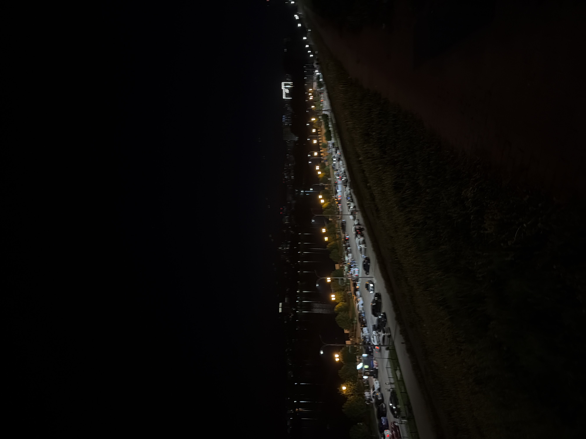 The image shows a nighttime scene in Izhevsk, Russia. On the right side of the frame, there is a paved road, lined with parked cars and streetlights. The cars appear to be parked alongside a grassy area, possibly a hill or slope. On the left side of the road, across from the parked cars, there is a body of water, likely a lake or river, visible under the dark sky. Distant lights reflect on the water's surface. In the distance, there are faint outlines of buildings and structures.