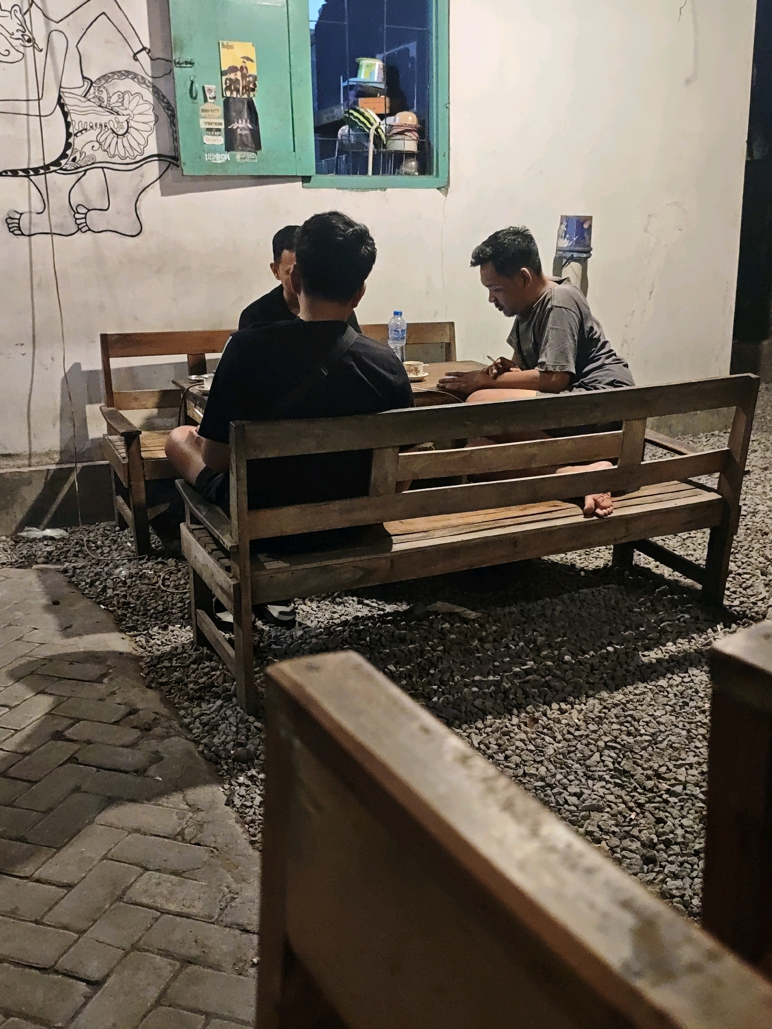 In the city of Ngasem, Indonesia, three individuals are seated around a wooden table in what appears to be an outdoor seating area. Two men are visible from the back, wearing dark tops, and are seated on a wooden bench facing the table. A third man, wearing a grey t-shirt, is seated on the opposite side of the table, facing away from the camera. He is holding a cigarette and appears to be engaged in conversation or activity at the table. The table has a bottle of water and a cup on it. The ground is covered in small, grey pebbles, and a tiled walkway is visible to the left. A white wall with a mural of a figure is present behind the individuals, and a green-framed window with items visible inside is above and to the right of the mural. The scene is dimly lit, suggesting it is evening or nighttime.