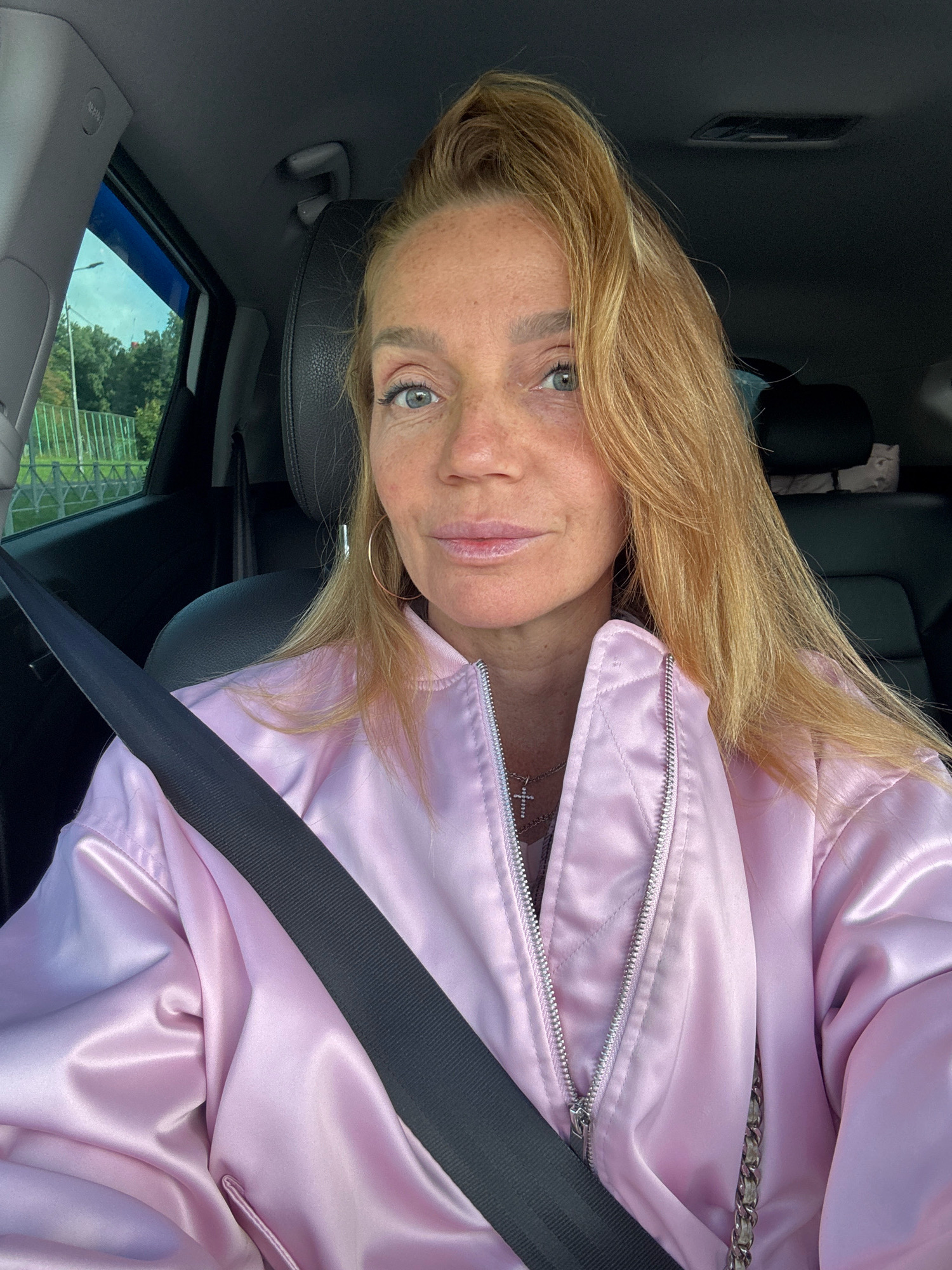 A woman with blonde hair and freckles, wearing a pink satin bomber jacket and a seatbelt, is in the driver's seat of a car. She is looking at the camera and her mouth is slightly open. The car's interior is visible, including the headrest of the passenger seat. Outside the car window, trees and a fence can be seen. The scene appears to be a daytime shot, possibly taken on a street or in a parking lot in an urban or suburban setting.