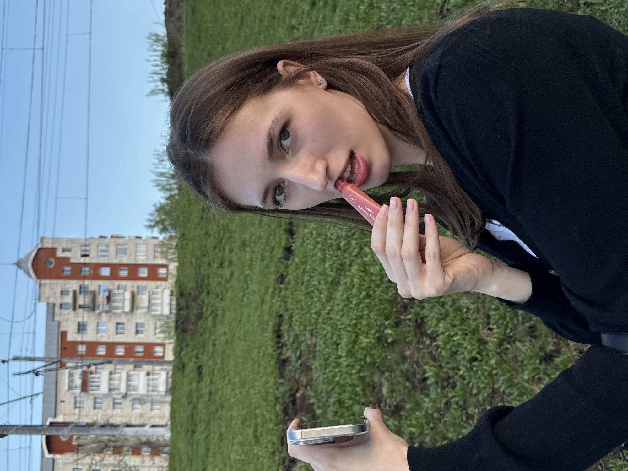 A young woman applies lip balm in Perm, Russia. She is fair-skinned with long brown hair, wearing a black cardigan over a white shirt. She holds a tube of lip balm to her lips with her right hand and looks towards the viewer. Her left hand holds a smartphone. In the background is a green grassy area, a tall residential building of Soviet-era architecture with light beige walls and red brick accents, and electrical wires stretching across the sky.