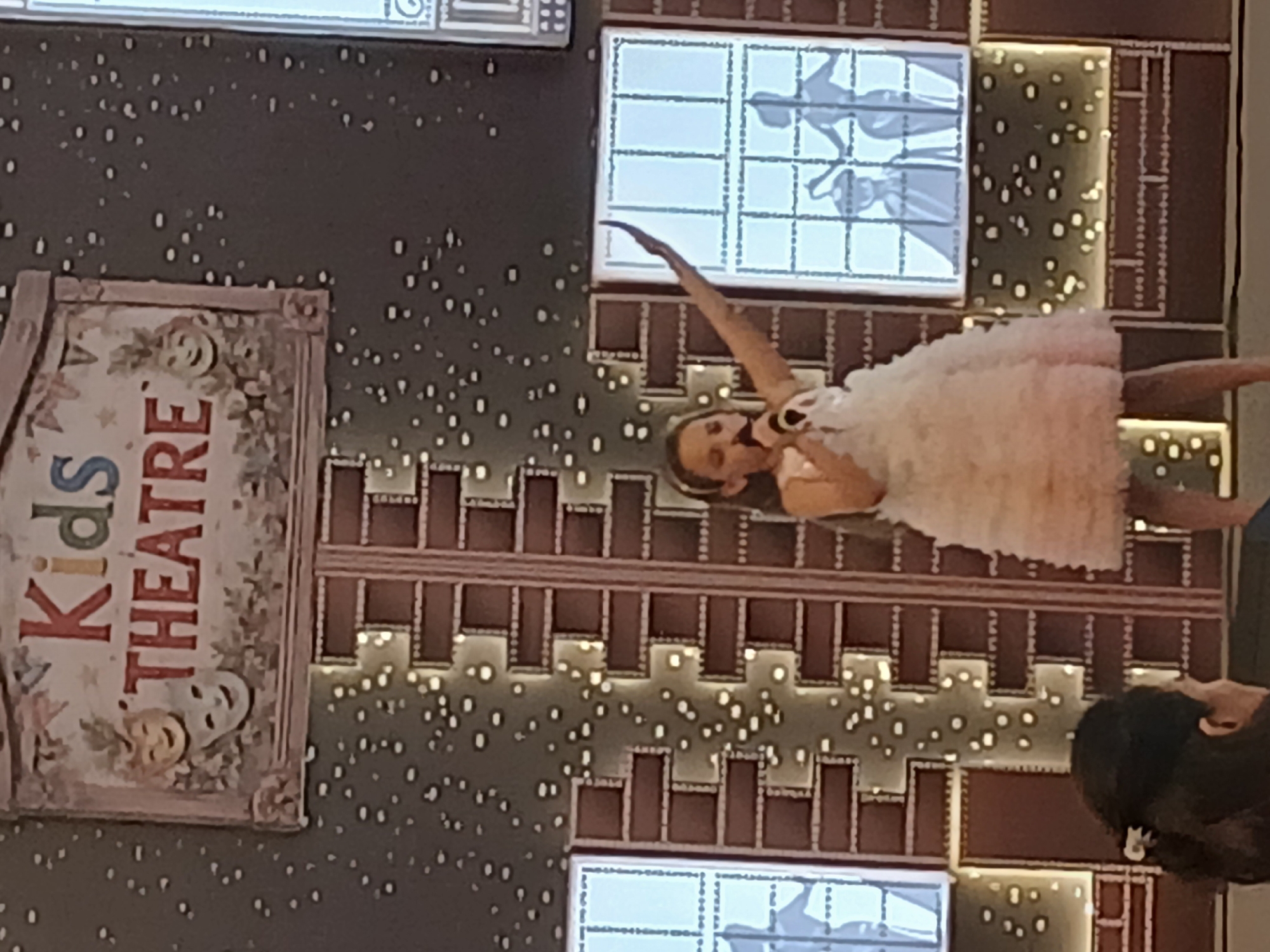 The image depicts a young girl in a white, frilly dress performing on a stage. She is holding a microphone and appears to be singing or speaking. Her arm is raised, and her body is angled towards the left of the frame. Behind her, the backdrop features illuminated panels with silhouettes of dancers, set against a brick-like pattern. The overall scene suggests a theatrical performance or entertainment event, likely for children, given the "Kids THEATRE" sign visible on the left. The sign itself is decorated with theatrical masks and floral motifs. The background is dark and speckled with small, white lights, creating a starry effect. The lighting on the stage is bright, highlighting the performer. The image is captured in portrait orientation and is slightly tilted.