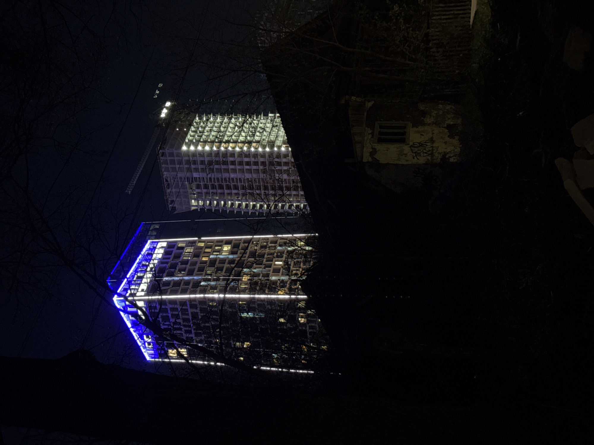 The photograph depicts two tall buildings at night, partially obscured by trees and foliage. One building is brightly lit with white lights, with a crane visible near the top. The other building is illuminated with white and blue lights. A smaller, older, weathered structure with a window is partially visible on the right side of the frame. The location is identified as the city of Izhevsk, Russia. No people or actions are clearly visible in the image.