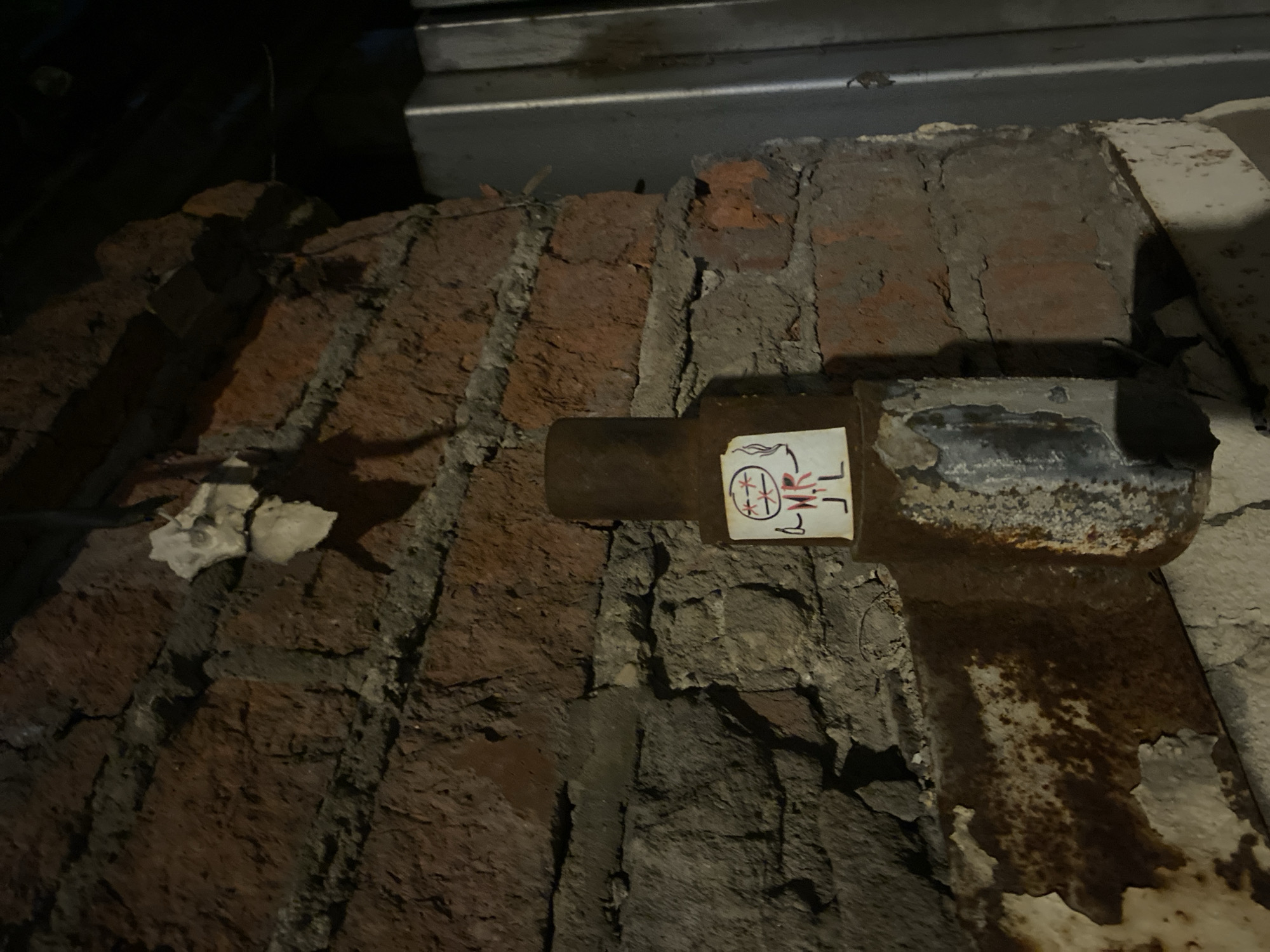 The image shows a section of exposed brick and crumbling concrete wall with a metal pipe protruding from it. A small sticker with red and black symbols is affixed to the pipe near the wall. The environment is dark and appears to be in a state of disrepair. The location is possibly in Moscow, Russia, based on the sticker's symbols.