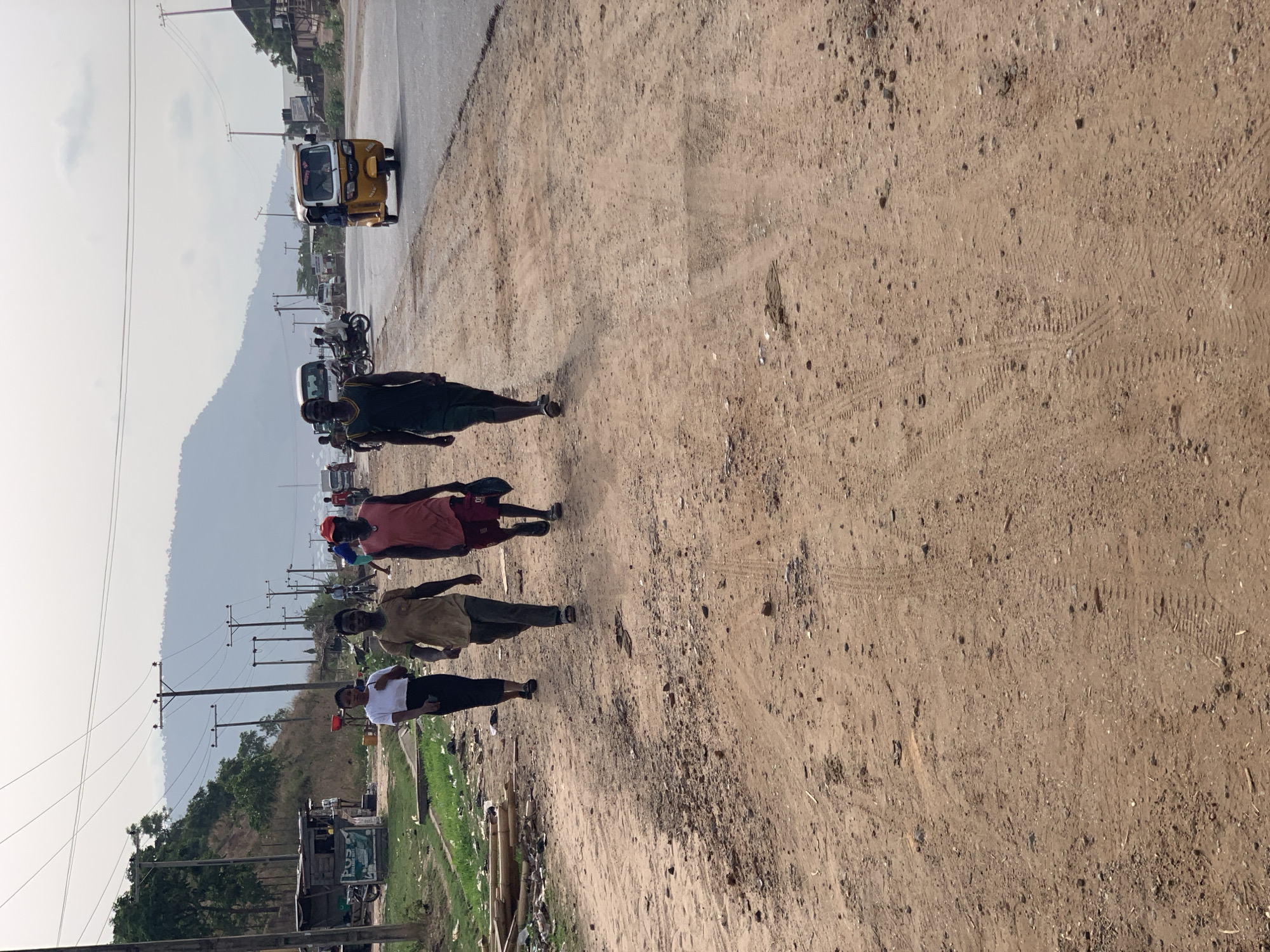 The image captures a daytime scene along a bustling road in what appears to be a semi-urban or rural environment, consistent with Lokoja, Nigeria. The photograph, when correctly oriented, shows a wide, dusty, unpaved dirt path on the right, running parallel to a paved road.
In the foreground, four men are walking along the dirt path, moving away from the camera. The closest man wears a white t-shirt and dark pants. Behind him are a man in a light brown shirt, another in a red t-shirt, and the furthest in a dark green or black t-shirt. The unpaved ground shows visible tire tracks and footprints.
On the paved road, a yellow three-wheeled vehicle (commonly known as a "keke napep" or auto-rickshaw in Nigeria) is prominently featured, also moving away from the viewer. Several motorcycles and other vehicles are visible further down the road.
The left side of the road is lined with wooden utility poles carrying power lines, extending into the distance. Below these poles, there is sparse vegetation, scattered debris, and some rudimentary structures or stalls. The background reveals a landscape of hazy hills or mountains under a bright, possibly overcast, sky.
The scene depicts daily life and movement, with people commuting on foot and vehicles on the road. The lighting suggests it is mid-day. No legible text is visible on any signs or objects.