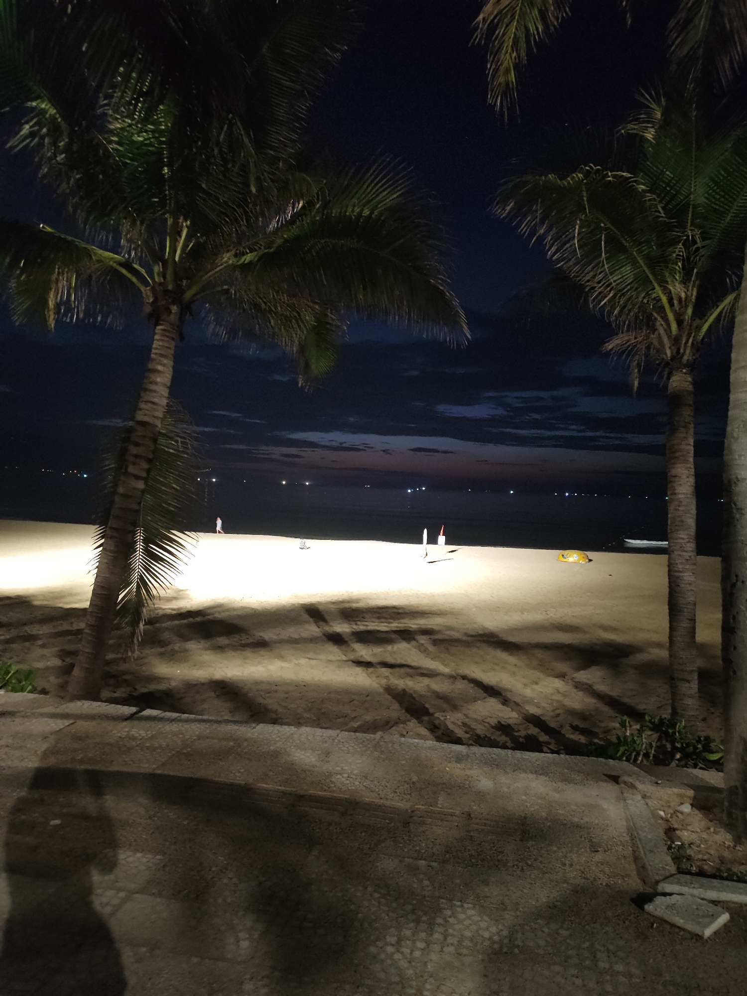 The image depicts a nighttime coastal scene in Đà Nẵng, Vietnam, viewed from an elevated position behind a concrete structure, possibly steps or a pathway. In the immediate foreground, a dark, indistinct shadow of a person, likely the photographer, is cast upon the light-colored concrete surface, which shows signs of wear and pebbled texture.
Several tall palm trees frame the composition on both the left and right sides, their dark trunks extending vertically and their fronds silhouetted against the dark sky, with some green foliage subtly illuminated by ambient light. Their elongated shadows are cast across the brightly lit sandy beach in the midground.
The wide expanse of the beach is illuminated by artificial light, appearing light yellow to white. A lone individual, dressed in light-colored clothing, walks leftward on the sand near the water's edge. To the right of the central axis, two white and red markers or flags are planted in the sand. Further to the right, a yellow, possibly inflatable, object lies on the sand, and a small, light-colored boat is partially visible, pulled ashore near the right edge of the frame.
The background features the dark, calm ocean. Across the distant horizon, numerous small, bright white lights are visible, indicating either distant boats or lights from the far coastline. The sky above is a deep indigo, with a faint reddish-orange glow along the horizon line, suggesting either very late dusk or very early dawn. Scattered dark cloud formations are present in the sky.