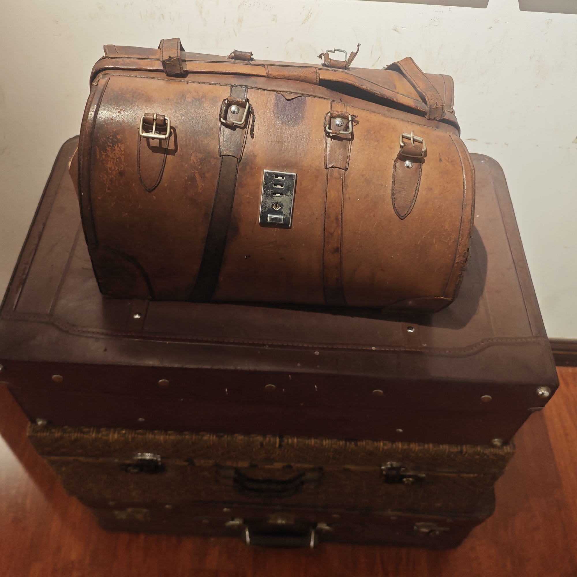 The image depicts a stack of three vintage suitcases on a wooden floor. The top suitcase is a brown leather duffel bag with metal buckles and straps. A metal combination lock is visible on the front of the bag. Below it is a dark brown suitcase with metal studs. At the bottom is another suitcase with a woven pattern and two handles visible. The background is a white wall. The photograph was likely taken indoors in an unspecified location in the Yuzhong District, China. No people or actions are present in the frame.