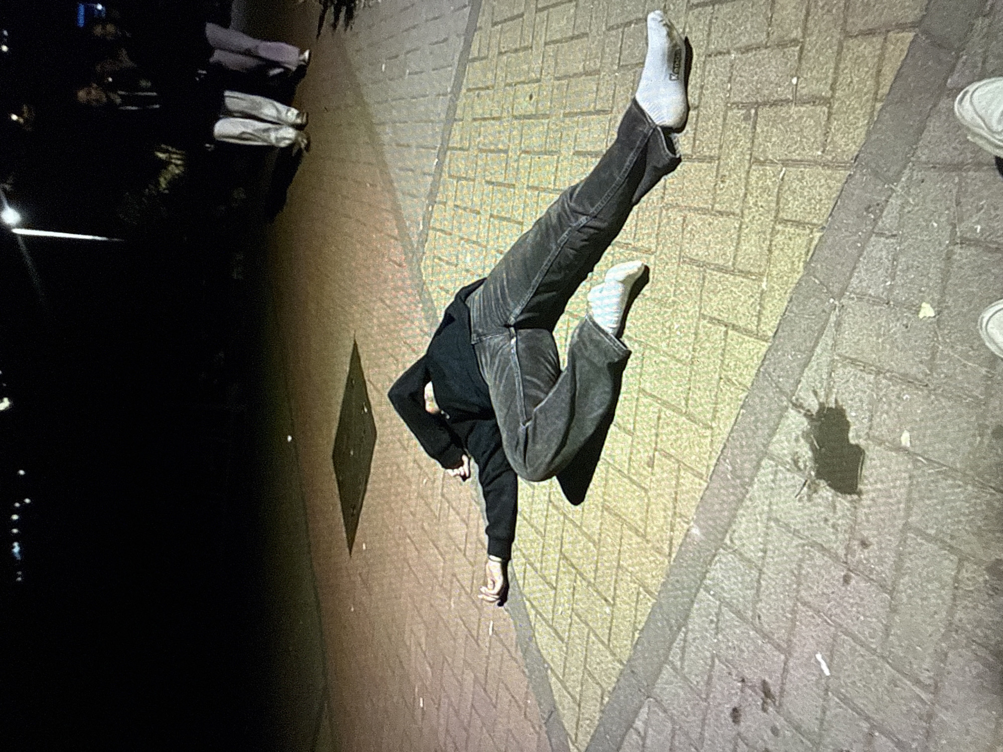 The image displays an outdoor nighttime scene on a paved surface in Kardzhali, Bulgaria. A single individual lies supine, slightly on their left side, on a brick paver walkway, extending diagonally across the midground. The person is clad in dark-colored jeans and a dark long-sleeved top with the hood pulled over their head, obscuring the face. Both feet are bare of shoes, wearing white socks; the sock on the right foot bears visible branding text, "Kappa." The left arm is extended towards the left side of the frame with the palm open and fingers slightly flexed, while the right arm is bent at the elbow with the hand positioned near the obscured head.
In the background, to the right, two indistinct individuals are standing upright on the same paved surface, partially illuminated by an unseen artificial light source. One person appears to be wearing light-colored trousers and a dark top, and the other wears darker trousers and a dark top. A square metal utility cover is visible on the pavement behind the prone individual.
On the grey-toned section of the pavement in the lower-left foreground, an irregular dark liquid stain or wet patch is present. Small white specks are scattered near the prone person's extended left hand. Long shadows are cast across the pavement, indicating the presence of overhead artificial lighting. The extreme bottom edge of the frame reveals the tips of two light-colored shoes, indicating the photographer's position. The background is largely dark, with blurred points of light suggesting distant urban illumination.