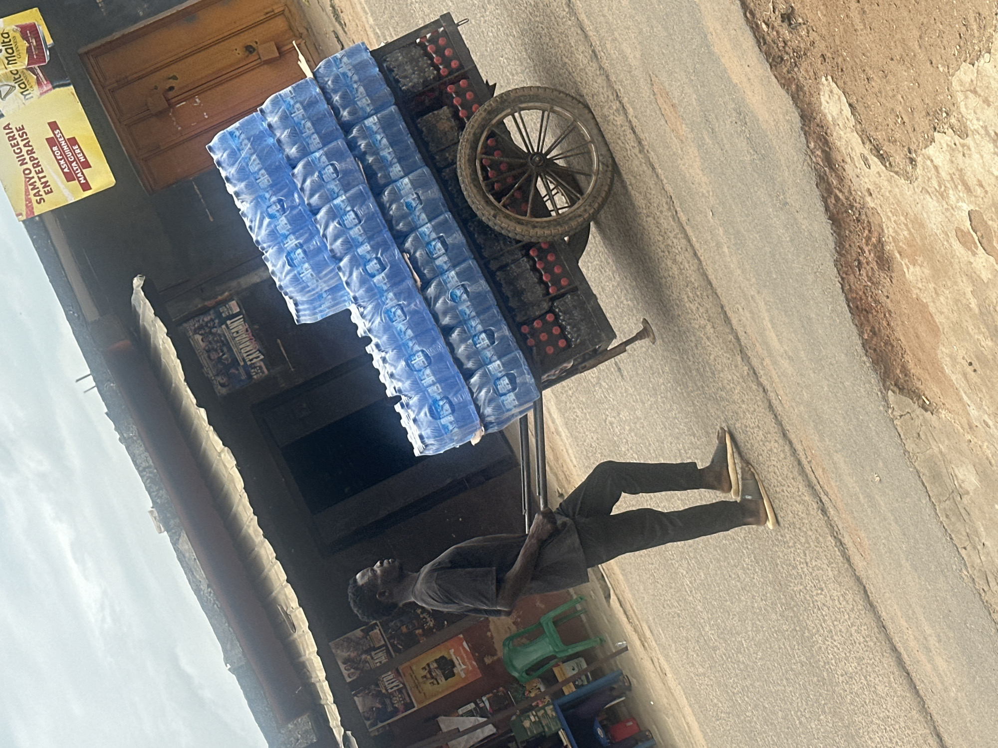 The image captures a male pushing a heavily loaded, two-wheeled handcart along a paved road in Jos, Nigeria, under an overcast sky.
The central figure is an adult male with dark skin and short dark hair, seen in profile, facing right. He wears a plain dark t-shirt, dark trousers, and light-colored open-toed sandals. His arms are bent, gripping the handles of the cart, indicating he is actively pushing it forward.
The handcart is constructed of dark-colored metal and features large, wooden-spoked wheels with metal rims, one of which is prominently visible on the right side. The cart is densely packed with merchandise. The upper two-thirds are stacked with multiple clear plastic shrink-wrapped packs of small plastic water bottles, each bottle having a blue cap. These packs are arranged horizontally in at least three layers. The bottom section of the cart holds numerous smaller bottles with red caps, contained within what appear to be black crates or carriers. A metal support leg extends from the front underside of the cart.
The ground beneath the cart and person is a grey, textured asphalt road. To the right of the road, an unpaved shoulder of dirt and gravel is visible. In the background, on the left, a dark building facade displays several posters, including one partially legible as "EXTRAVAGANT." A bright green plastic chair and a partial blue table are positioned near this building. Further to the right, another building features a prominent yellow rectangular sign that reads "SAMYO NIGERIA ENTERPRISE" and "ASK FOR MALTA GUINNESS HERE" in red text. Adjacent to this sign, two red and white "Malta Guinness" advertisements are displayed, and a dark brown wooden door with metal hinges is visible. The sky above is uniformly light grey, indicating overcast conditions. A concrete ledge or roofline is visible above the buildings.