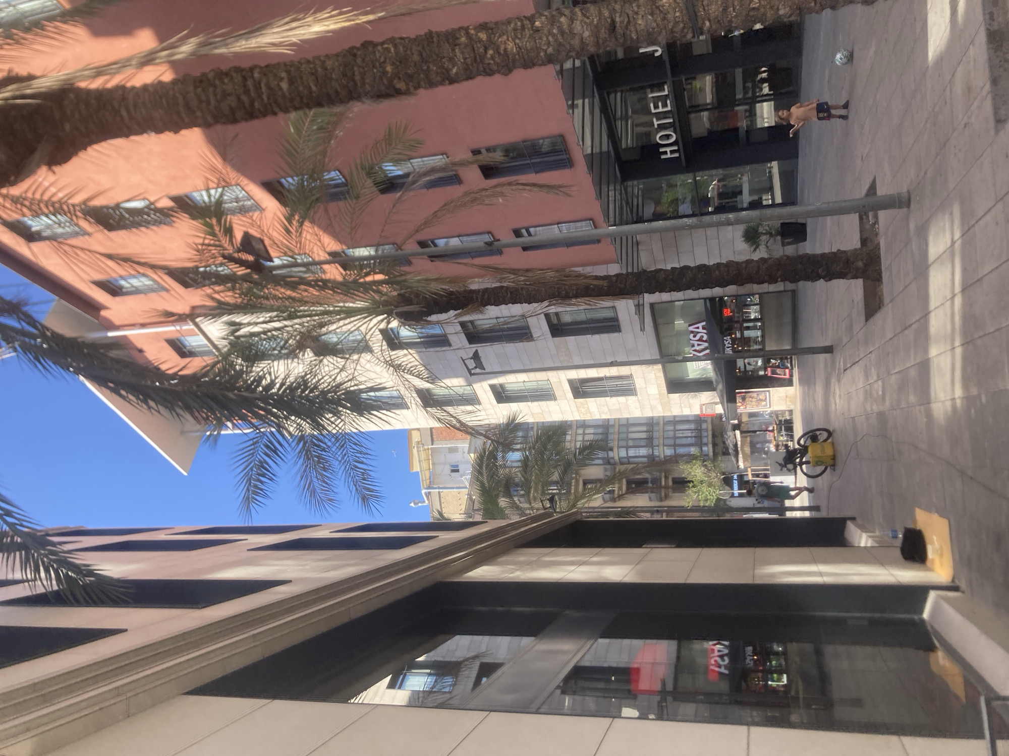 The image depicts a street scene in Barcelona, Spain, with a focus on the architecture and urban environment. On the left, a modern building with large glass windows and beige facade dominates the foreground, reflecting the surrounding structures and sky. Across the street, a salmon-colored building stands out, featuring multiple windows and balconies, with a prominent "HOTEL J" sign visible near its entrance. Palm trees are interspersed along the sidewalk, casting shadows on the paved ground. In the middle ground, a yellow delivery bag is attached to a bicycle, suggesting a commercial or delivery service. Further down the street, a young child is playing with a soccer ball on the sidewalk. The overall scene is bathed in sunlight under a clear blue sky, indicating a warm day. The perspective is low, looking up towards the buildings and trees.