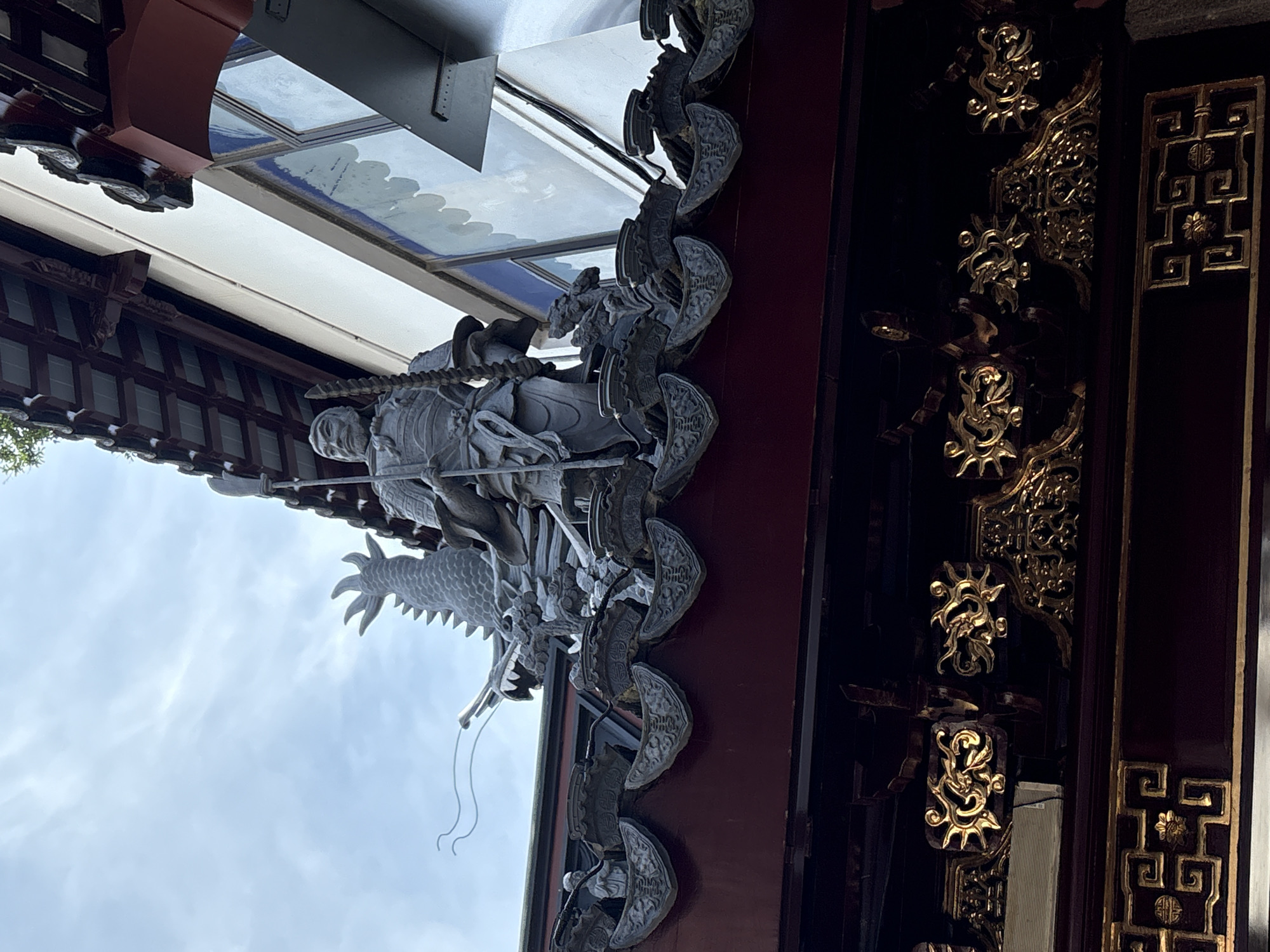This is a photograph taken from a low angle, looking up at the roofline of a traditional Chinese building in Shanghai, China. The building features ornate, dark red eaves with intricate gold detailing. Prominently displayed on the roof is a large, grey statue of a warrior in armor, holding a spear. Behind the warrior, a detailed dragon sculpture with sharp fins and flowing whiskers is visible. The sky above is overcast with pale clouds. The architectural style suggests a temple or historical structure, typical of areas like Yu Garden.