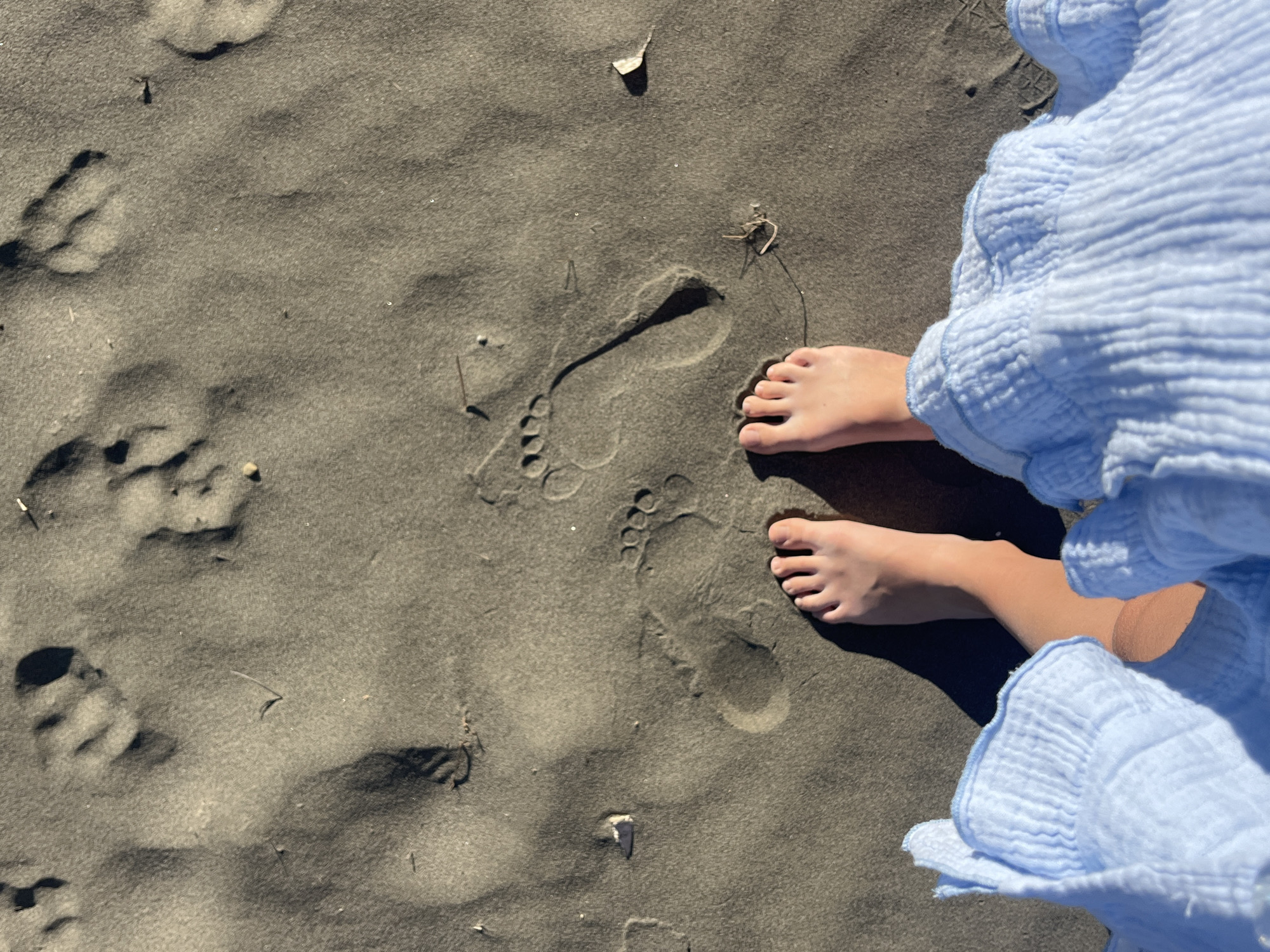 The image shows a pair of bare feet standing on dark, wet sand. Footprints are visible in the sand, indicating recent passage. A light blue, ruffled fabric is visible at the bottom of the frame, suggesting the person is wearing a dress or skirt. The scene appears to be a beach. The location is the city of Pistula, Montenegro.