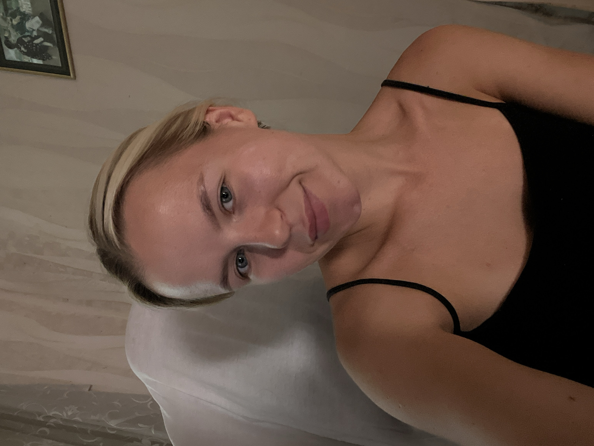 A young Caucasian woman with blonde hair pulled back is seen in a close-up, low-angle shot. She is wearing a black spaghetti-strap tank top and is seated on a light-colored, plush chair, partially covered by a white drape. Her head is tilted slightly to her right, and she is looking directly at the camera with a subtle smile. Her skin appears smooth and lightly flushed, with a faint glow, suggesting she might be wearing makeup or has a natural radiance. The background consists of a light beige wall with subtle vertical textured lines and a framed picture hanging on it. To the left, a light-colored, textured curtain is visible, creating a soft, diffused lighting effect. The overall impression is that of a candid, self-taken photograph in an indoor setting.