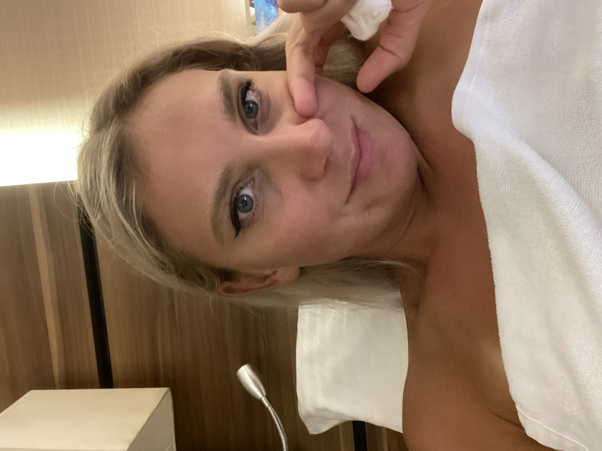A blonde woman is lying in bed, wrapped in a white towel. She is looking at the camera and touching her nose with her finger. The room has wooden walls and a white pillow. There is a small lamp attached to the wall above the pillow.