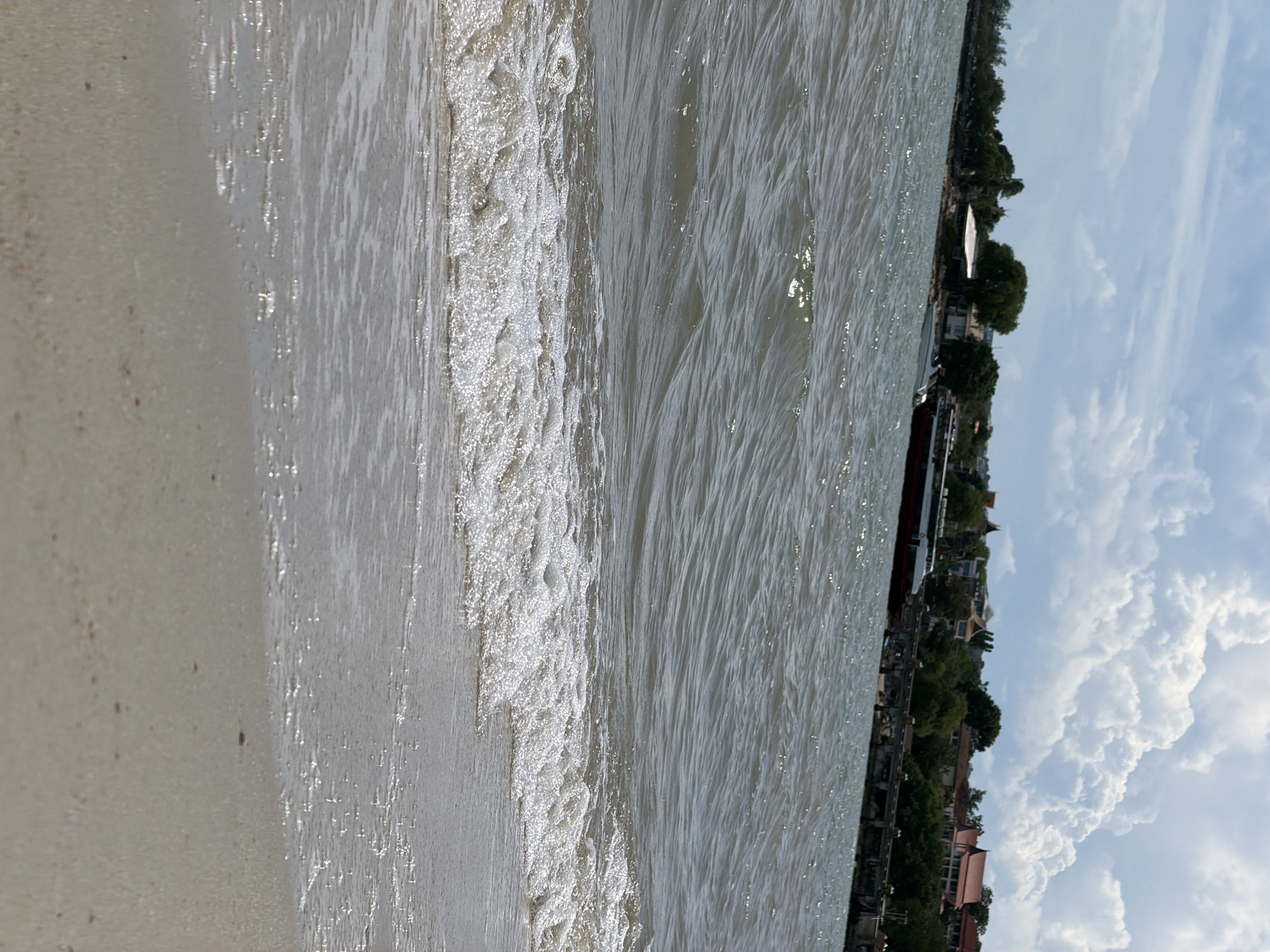 The image depicts a tranquil scene at the shore in Baan Bang Rak, Thailand. The foreground is dominated by a sandy beach with gentle waves lapping at the edge, creating a foamy, shimmering line. The water, a muted grey-green, shows the rippling effect of a moderate breeze. In the mid-ground, a long, red and white boat with multiple windows is docked or moored near a stone pier. Beyond the pier, lush green trees and a scattering of buildings with red-tiled roofs line the distant shore, suggesting a coastal town or village. The sky is a mix of light blue with large, fluffy white clouds, hinting at fair weather with potential for light rain. There are no people visible in the frame, and the overall impression is one of natural beauty and quietude.