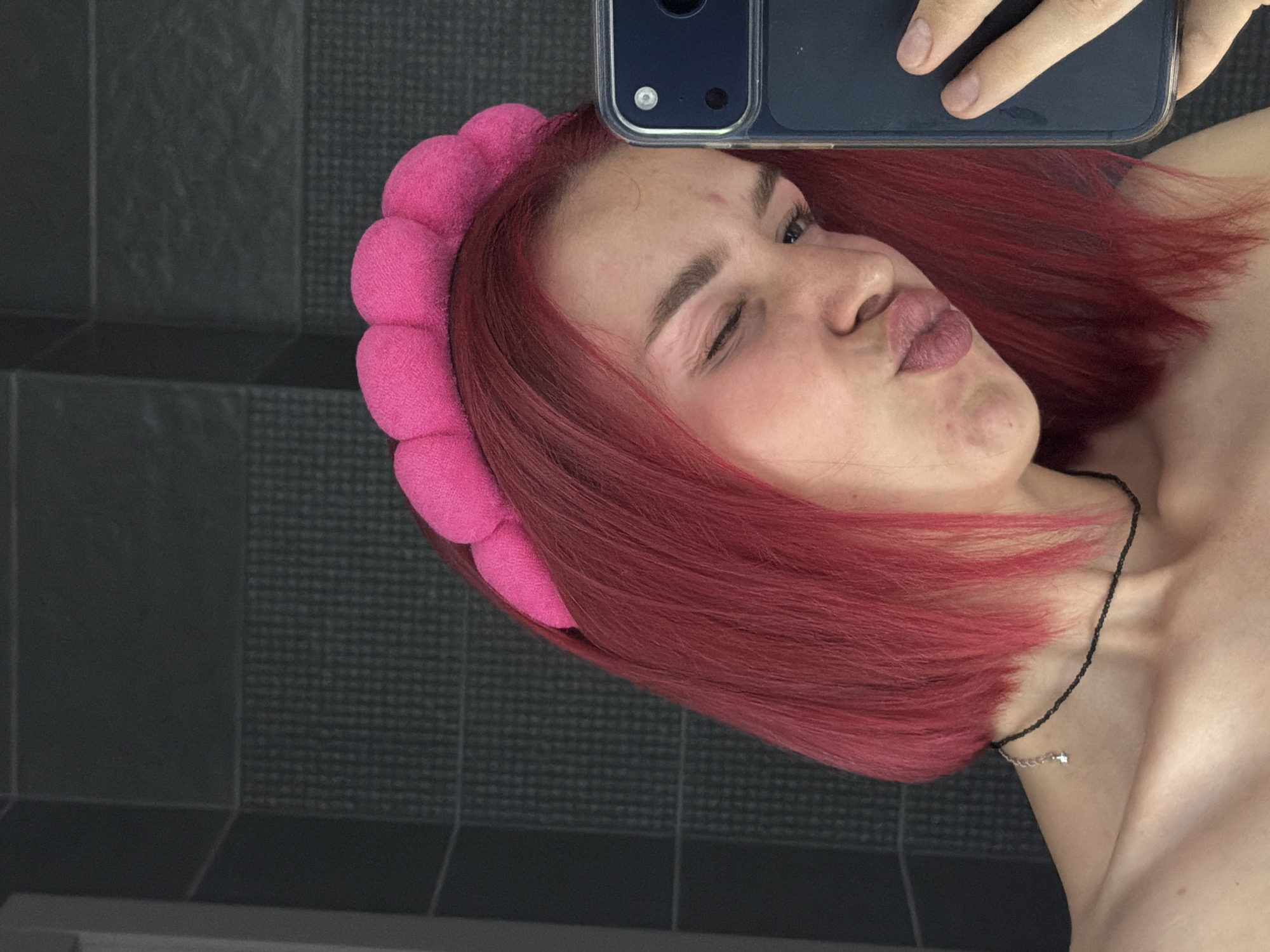The image depicts a close-up selfie of an individual in the city of Chaikovsky, Russia, taken in a room with dark gray tiled walls. The person has short, bright pink hair and wears a puffy, segmented pink headband. Their left eye is winked shut, and their lips are pursed in a pout. A thin, black beaded choker with a small silver charm adorns their neck, visible above bare shoulders. A dark smartphone in a clear case is held by the individual's right hand, partially obstructing the lower right side of the face. The background consists of large, dark gray rectangular wall tiles, with a lower section featuring smaller, textured dark gray tiles.