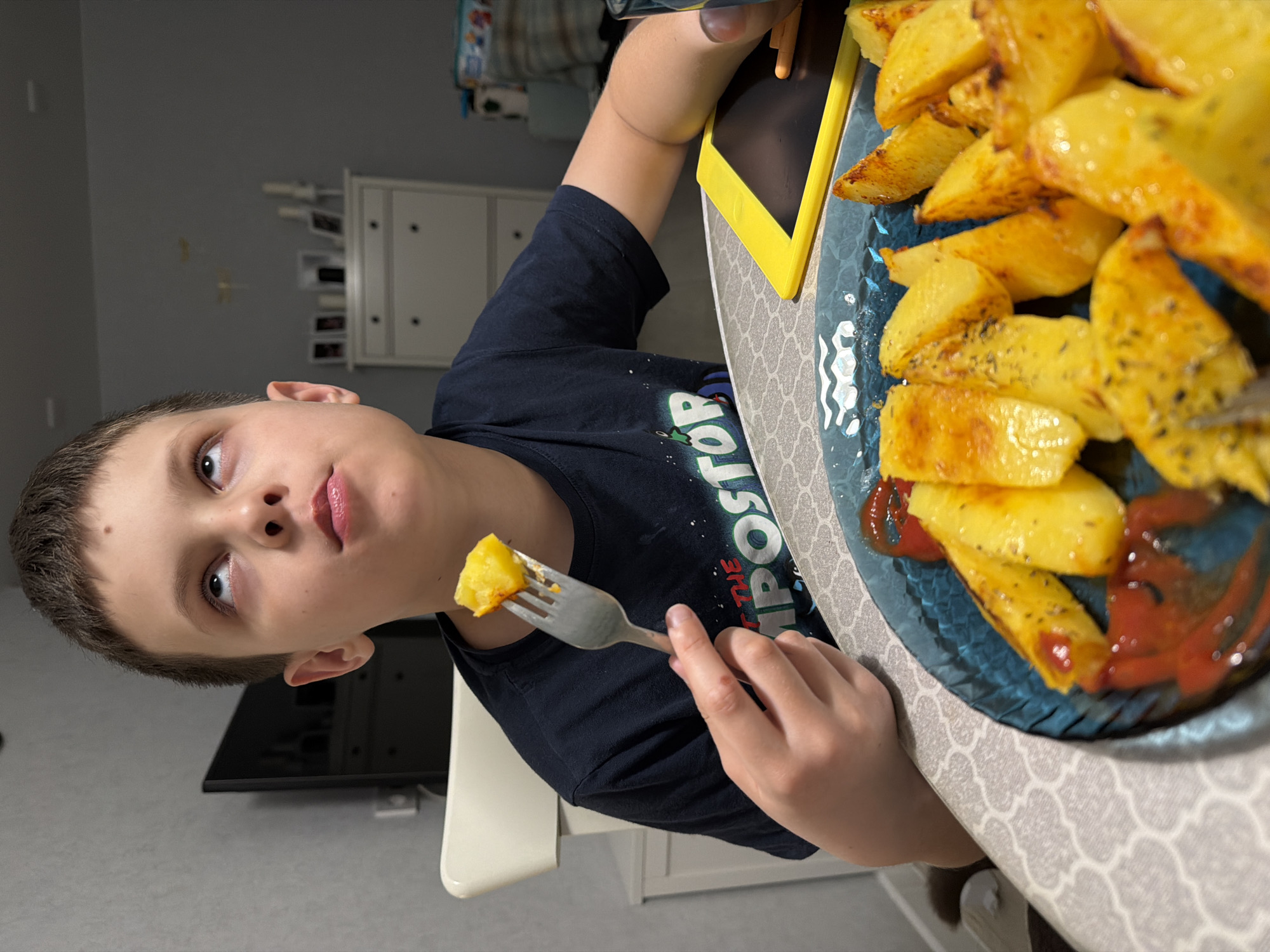 A young boy sits at a table in an indoor setting, possibly a home, eating baked potato wedges. He holds a piece of potato on a fork. A blue glass plate filled with more potato wedges and ketchup sits on the table in front of him, near a yellow tablet. The boy is wearing a dark blue t-shirt. Behind him, there's a television and a white cabinet. The table is covered with a patterned tablecloth. The exact location within Votkinsk, Russia, cannot be determined from the image.
