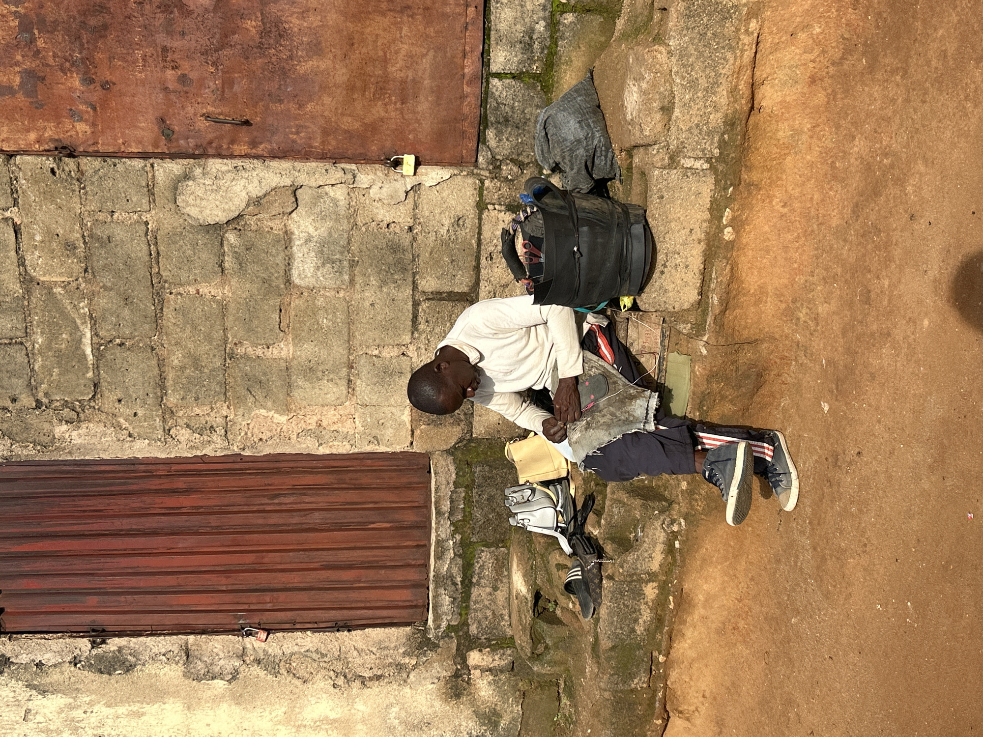 A man is sitting on stone steps outside a building, working on what appears to be a shoe. He is wearing a light-colored long-sleeved shirt, dark pants with red and white stripes on the side, and dark sneakers. In front of him on the steps are several items, including a black bucket containing tools, a gray bag, a yellow purse, and a white handbag, and a pair of black sandals. The building has a red metal door on the left and a rusty metal door on the right, both with padlocks. The wall is constructed of rough stone blocks. The ground in front of the steps is reddish-brown dirt. The scene suggests an outdoor workshop or workspace in a part of Jos, Nigeria, possibly a residential or commercial area characterized by its modest construction and direct engagement with street-level commerce or repair services.