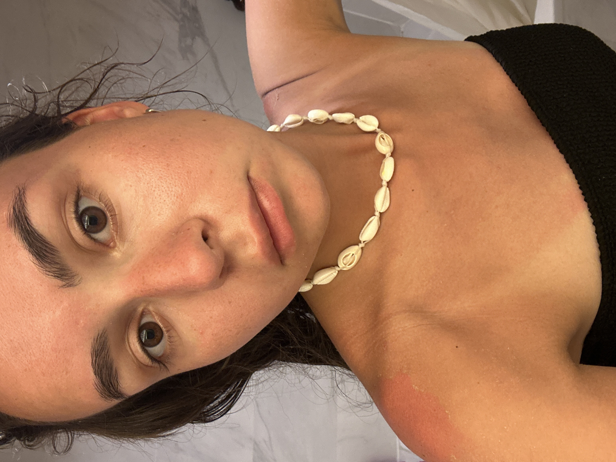 A close-up, angled view focuses on a woman. She wears a cowrie shell necklace and a black top. Her skin shows signs of sun exposure, particularly on her shoulder. The background is a blurry white, possibly a wall or tiled surface. There are no distinct features suggesting a location within Pattaya City, Thailand.