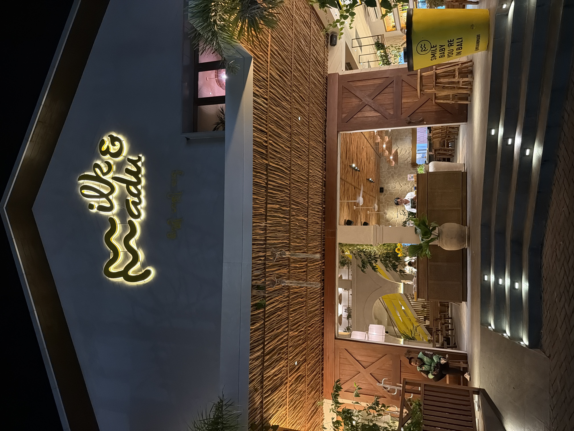 This media file captures the exterior of a modern, yet traditionally-styled building, likely a restaurant or cafe, at night in Kuta, Indonesia. The dominant feature on the left is a white A-frame section with a large, glowing yellow neon sign that spells "Silky Madu" in a flowing script, with smaller text "crafted bakery" visible beneath it.
To the right of this section, a significant portion of the wall is clad in natural, horizontal woven materials, giving it a rustic texture. Further right are large wooden double doors with an "X" motif, resembling barn doors, and large glass windows revealing the illuminated interior. Inside, warm lighting highlights wooden tables, chairs, and a counter where a person appears to be working. Potted plants and decorative lighting fixtures are also visible within.
On the far right, a wide staircase with embedded square lights leads upwards. Beside the stairs, a tall, yellow cylindrical sign prominently displays the text "SMILE BABY YOU'RE IN BALI" and "GULIHANKMADU". Below this sign, a person wearing a green jacket is seated on a wooden bench.
The scene is lit entirely by artificial lights, indicating it is nighttime. Palm fronds and other greenery add a tropical touch to the architecture. The overall impression is that of a welcoming, well-lit establishment open for business.