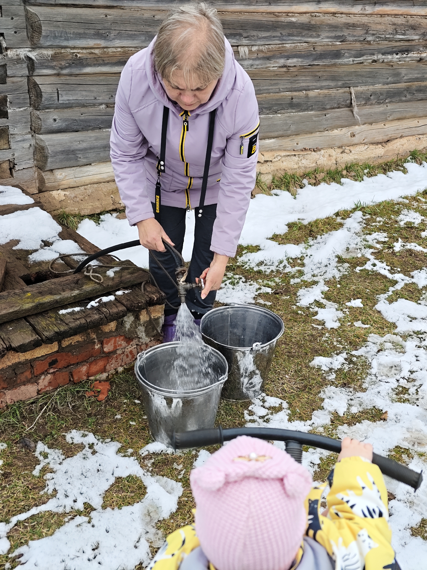 In Уральское, Russia, an elderly woman with short, light hair wearing a light purple jacket, dark pants, and purple boots is filling two metal buckets with water from an outdoor well. The well is constructed with bricks and wood. A black hose is connected to the well. The ground is covered with grass and patches of snow. In the foreground, a child wearing a pink hat and yellow jacket is partially visible, holding onto a black handlebar. A wooden building is visible in the background.