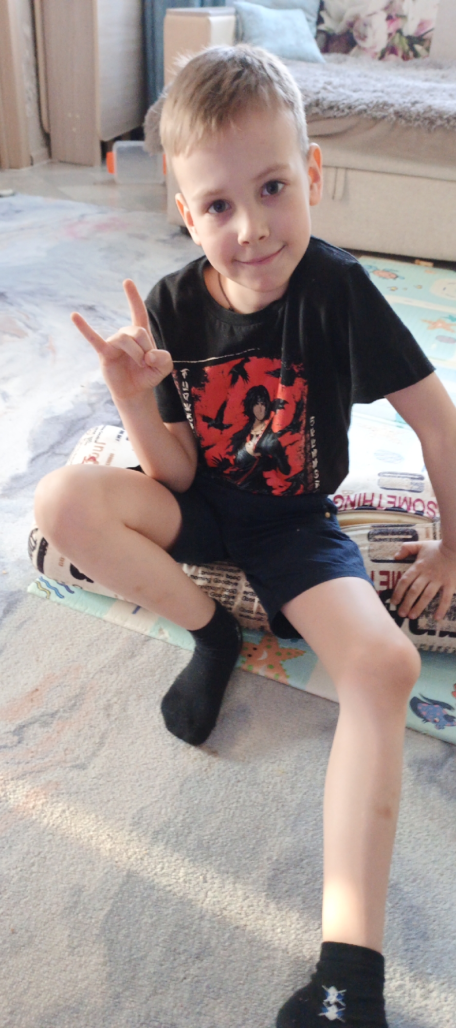 A young boy is seated indoors. He is wearing a black t-shirt with a red and black graphic design on the front, dark blue shorts, and black socks. He is making a hand gesture, specifically holding up his index and pinky fingers in a “rock on” or “devil horns” sign. He is sitting on a printed cushion. The background shows a carpeted floor and a glimpse of a bed with pillows. The location is indicated to be in the city of Chaikovsky, Russia.