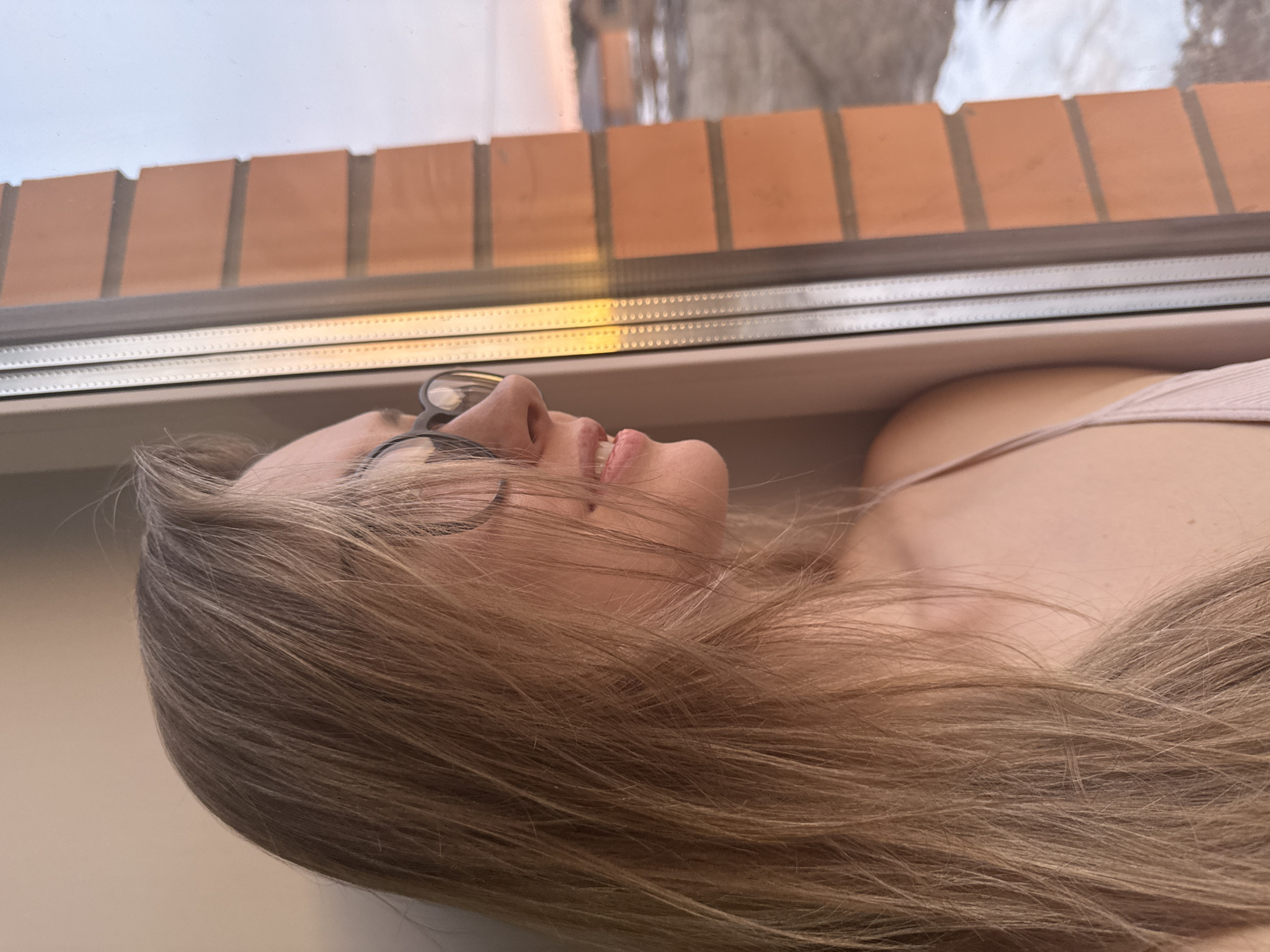 A light-skinned individual with long, blonde hair and wearing eyeglasses is resting their head on a window sill. The window is framed by a brick wall visible outside. The individual is wearing a light pink top. The location is Chaikovsky, Russia.